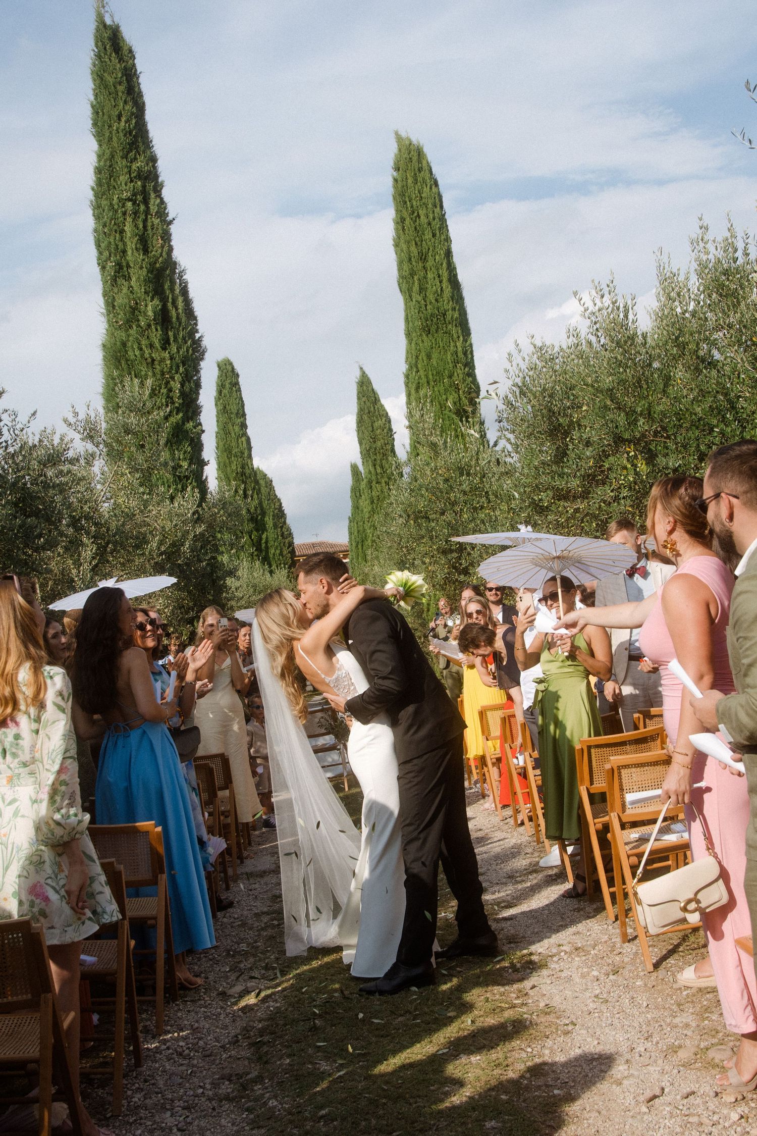 Joyful wedding celebration on path lined with cypress trees and white parasols.