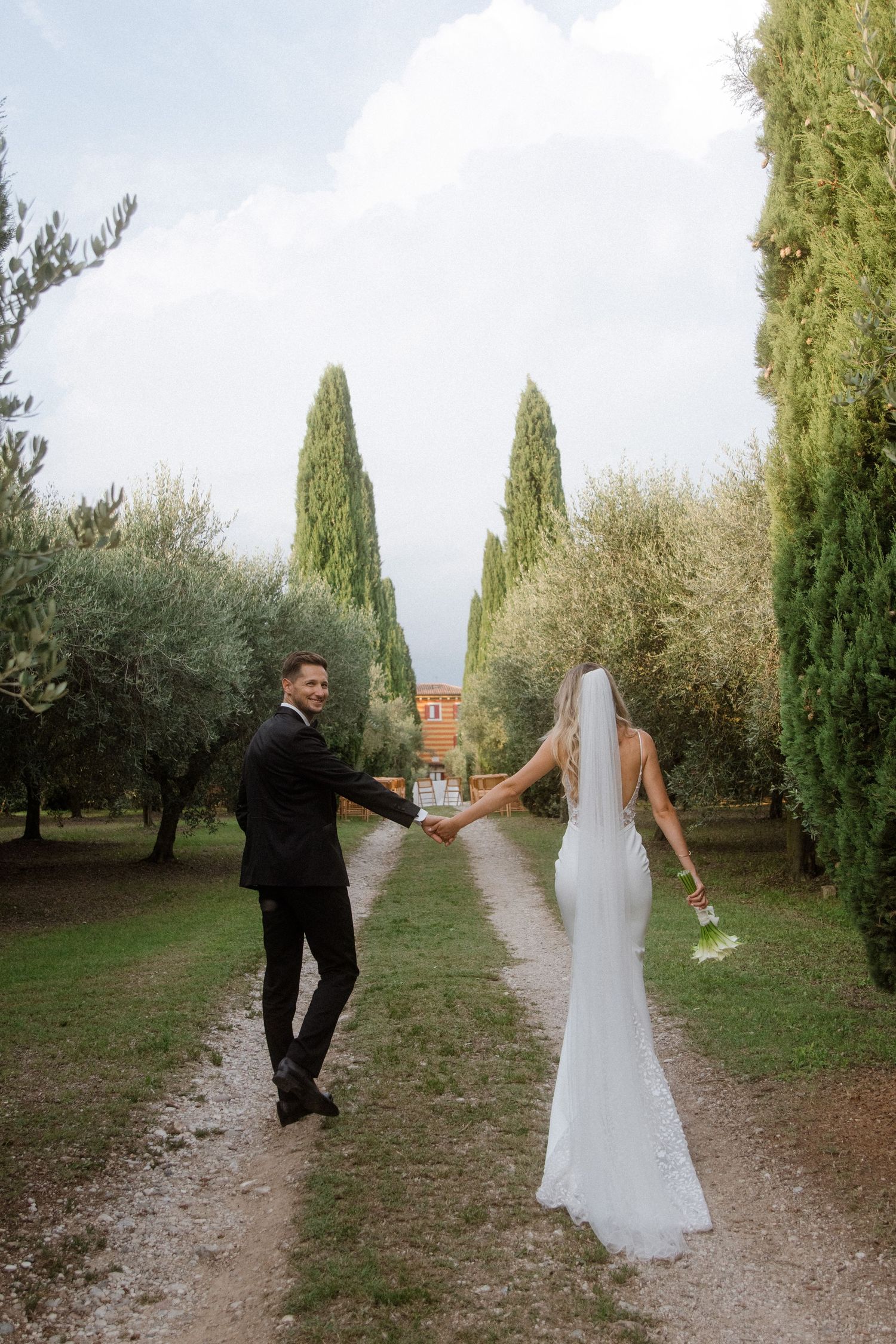 Wedding couple holding hands while walking along a scenic garden path lined with cypress trees.