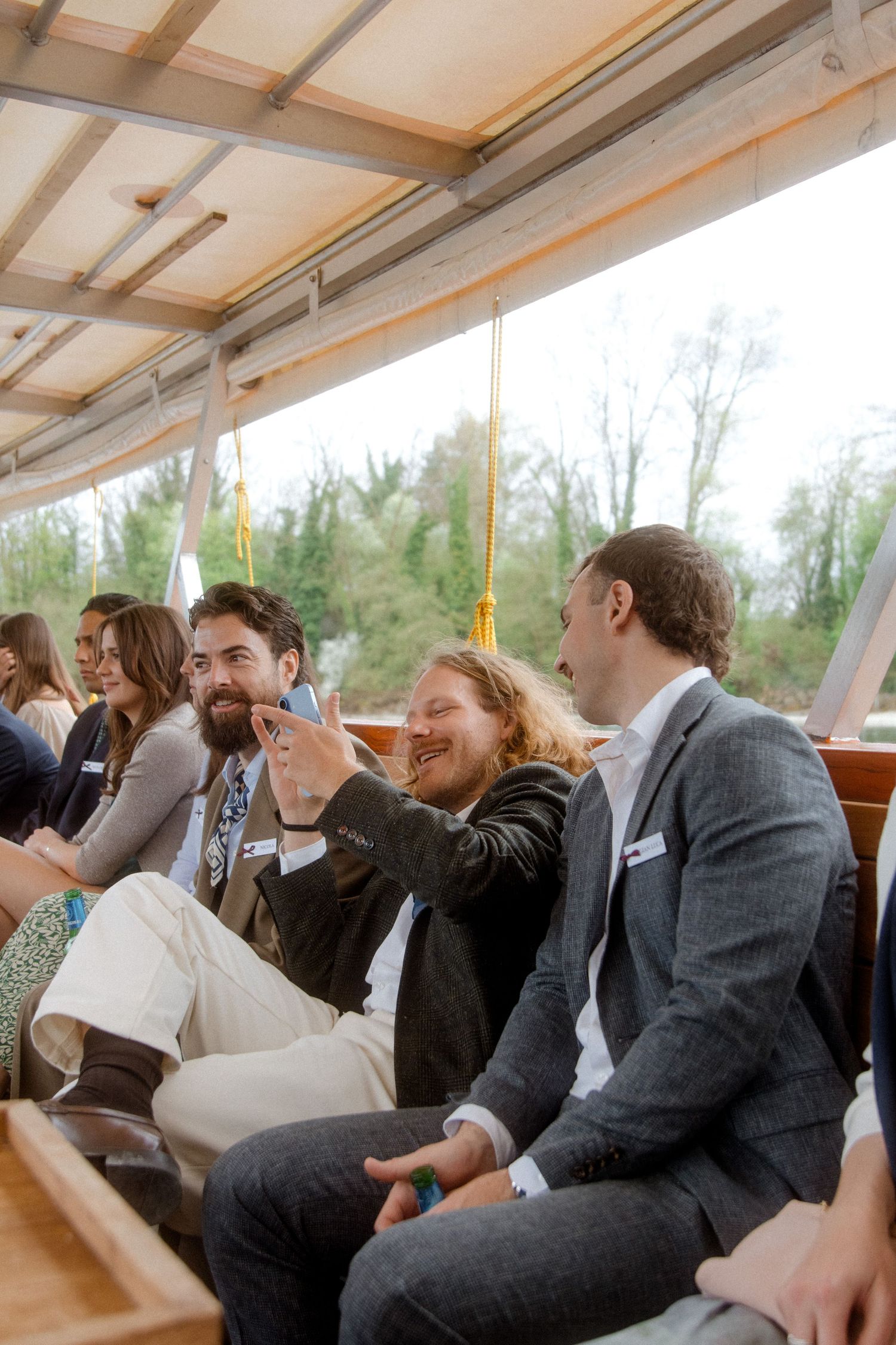 Men laughing and taking selfie on covered boat during wedding celebration.