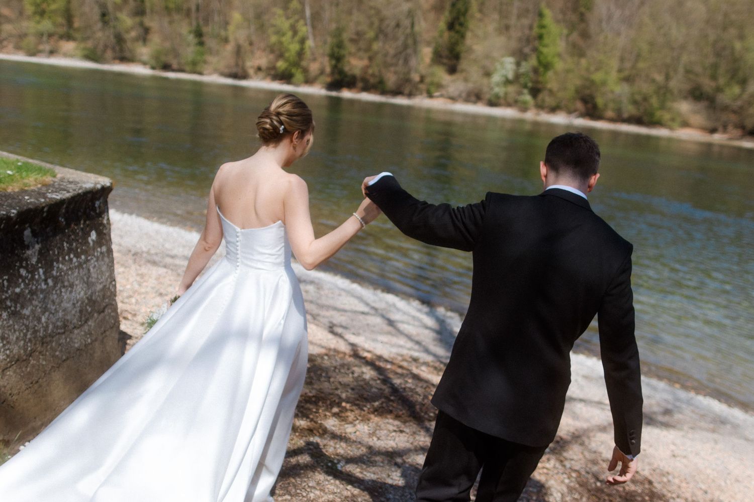 Bride and groom walking hand in hand along a scenic riverside on their wedding day.