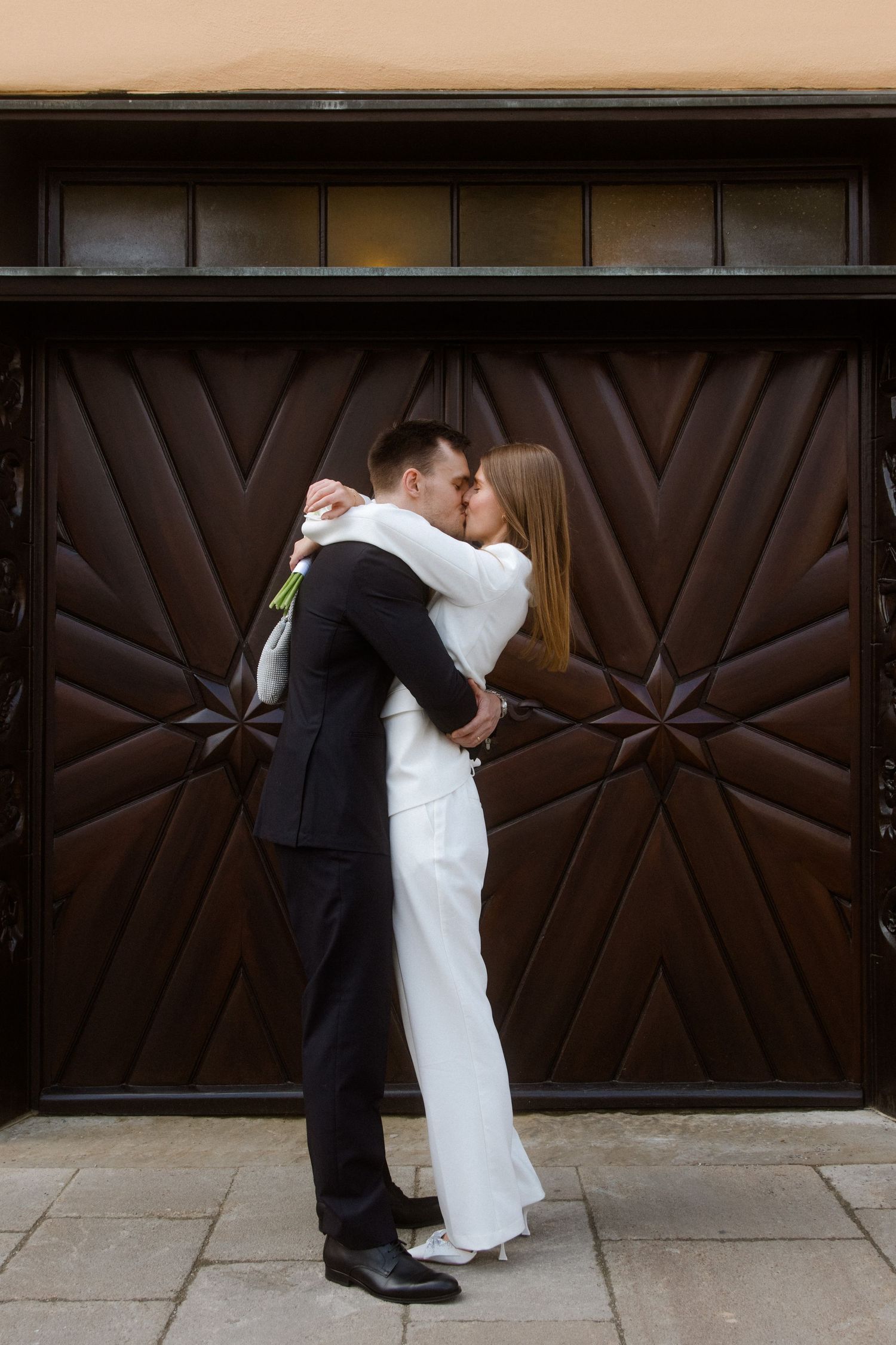 Newlyweds sharing a romantic kiss in front of ornate wooden doors on their wedding day.