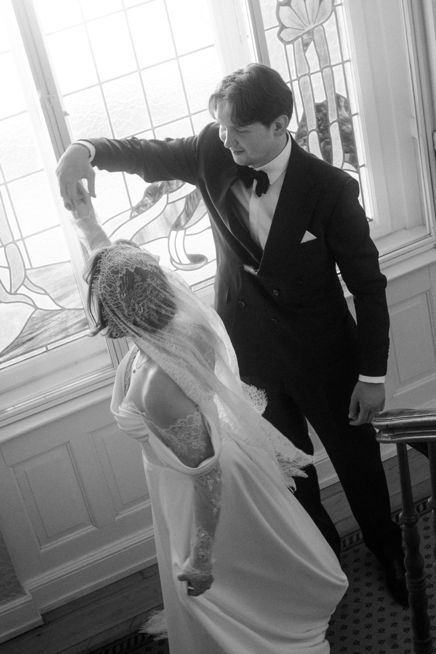 Black and white photo of a groom in a tuxedo spinning his bride in a wedding gown near a stained glass window.