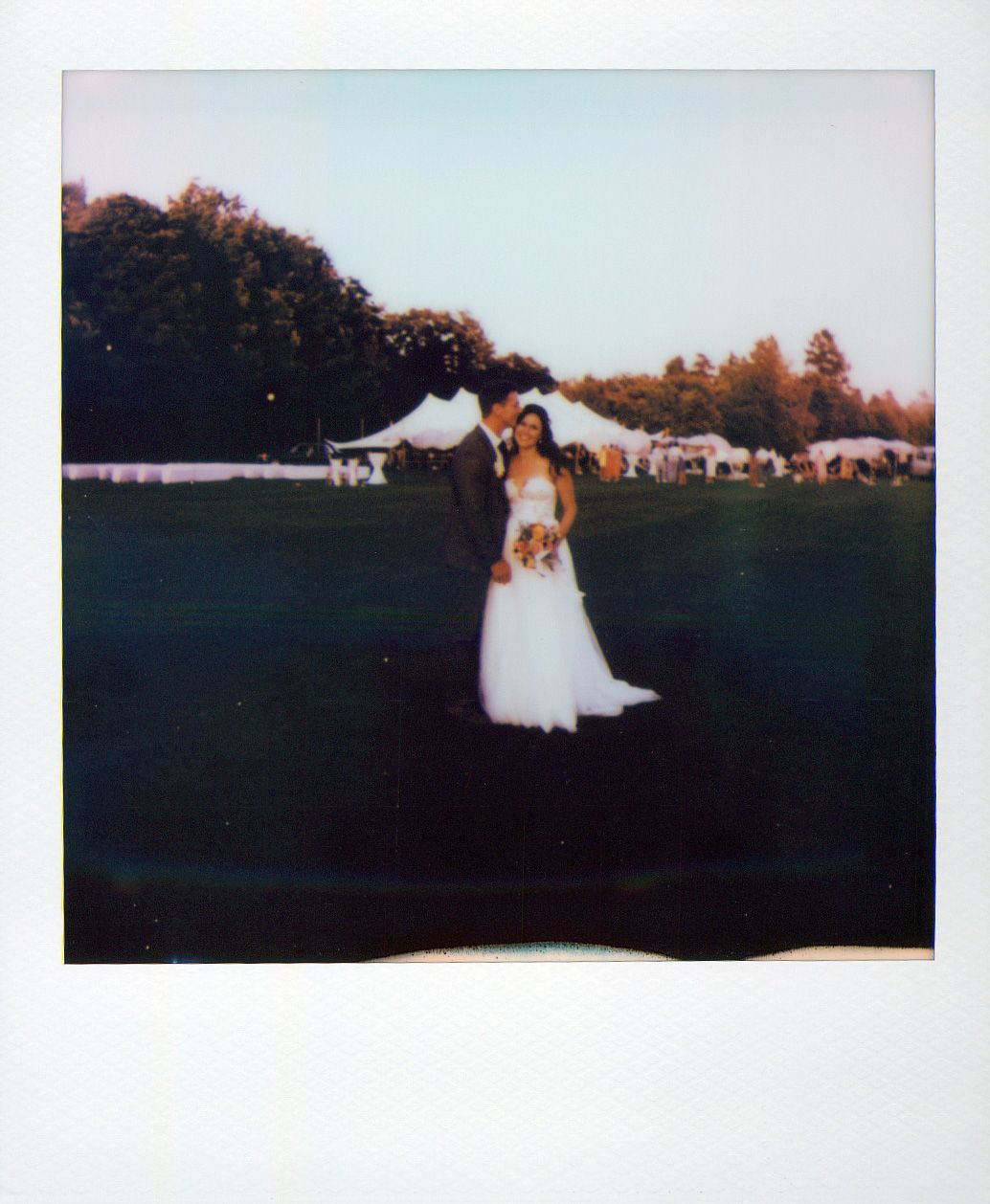A row of white event tents set up in a field at sunset with autumn trees in the background.