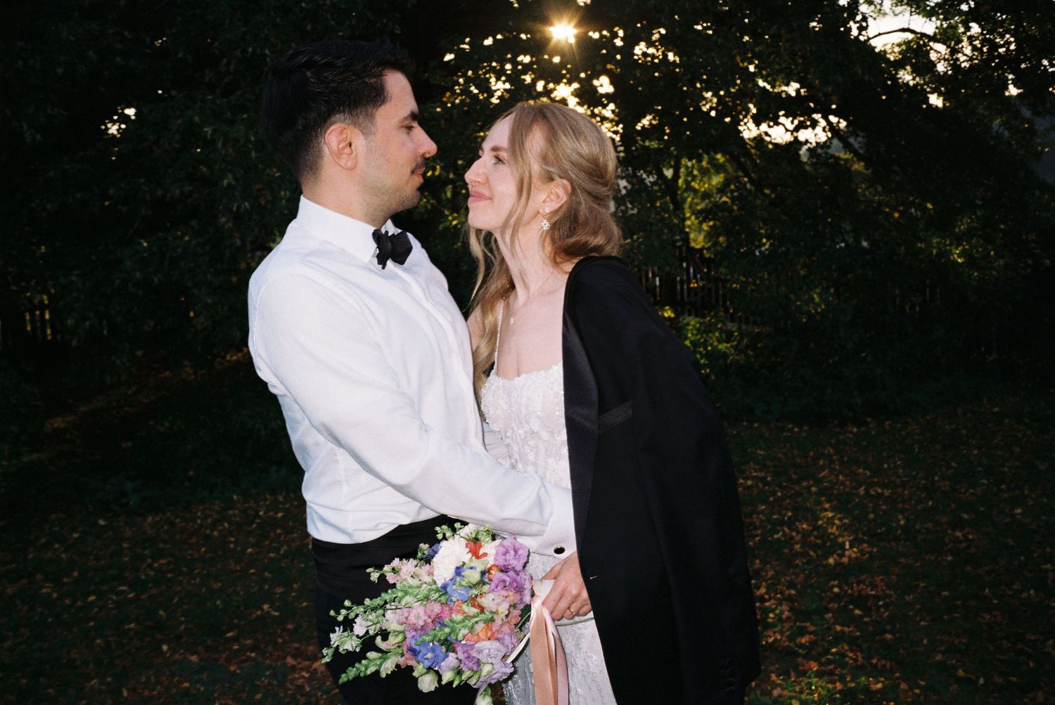 A romantic couple in wedding attire embraces outdoors at dusk with twinkling lights in the background.