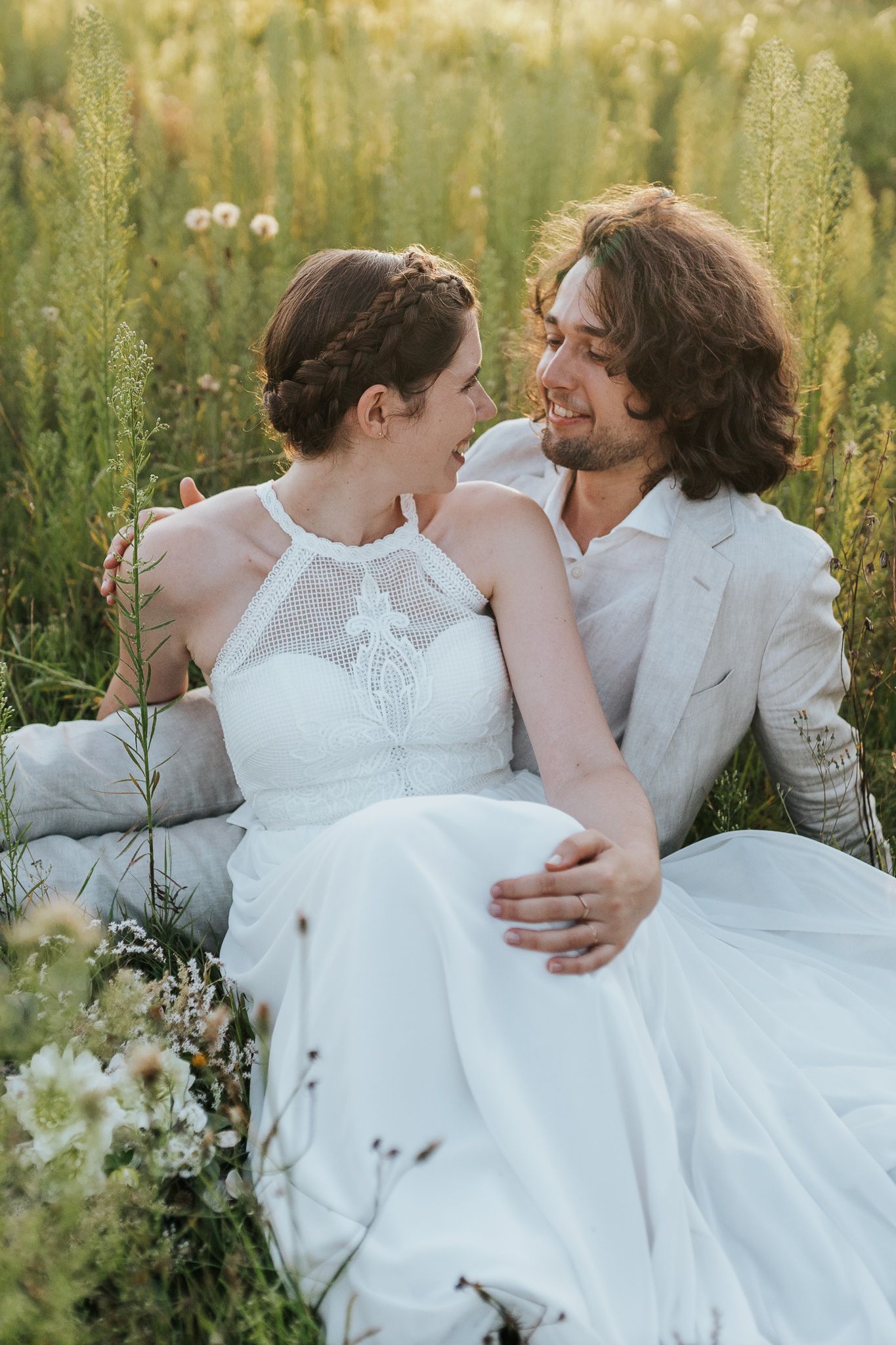 A romantic couple in white sits together in a sunlit meadow with wildflowers.