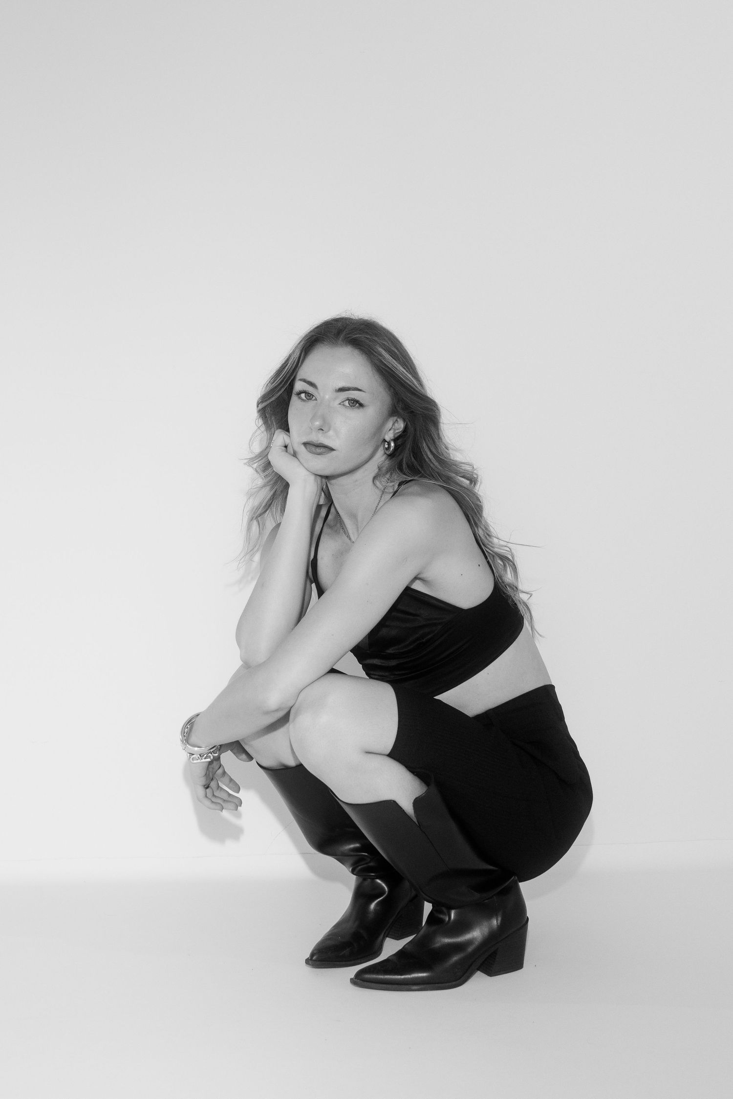 Black and white photo of a woman in a crop top, mini skirt, and boots, crouching gracefully in a studio setting.