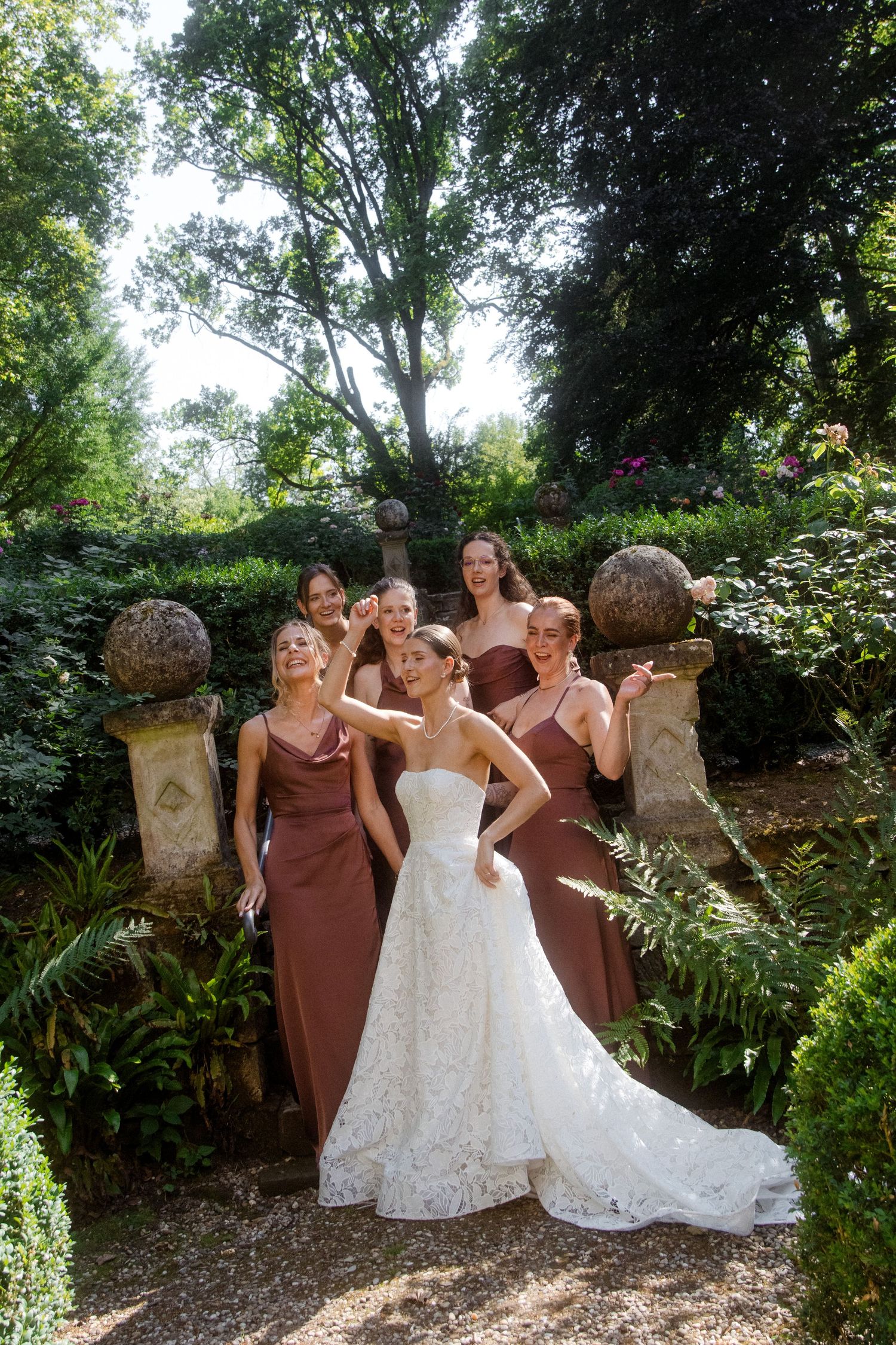 Bride in white lace gown poses with five bridesmaids in rust dresses in a lush garden setting.