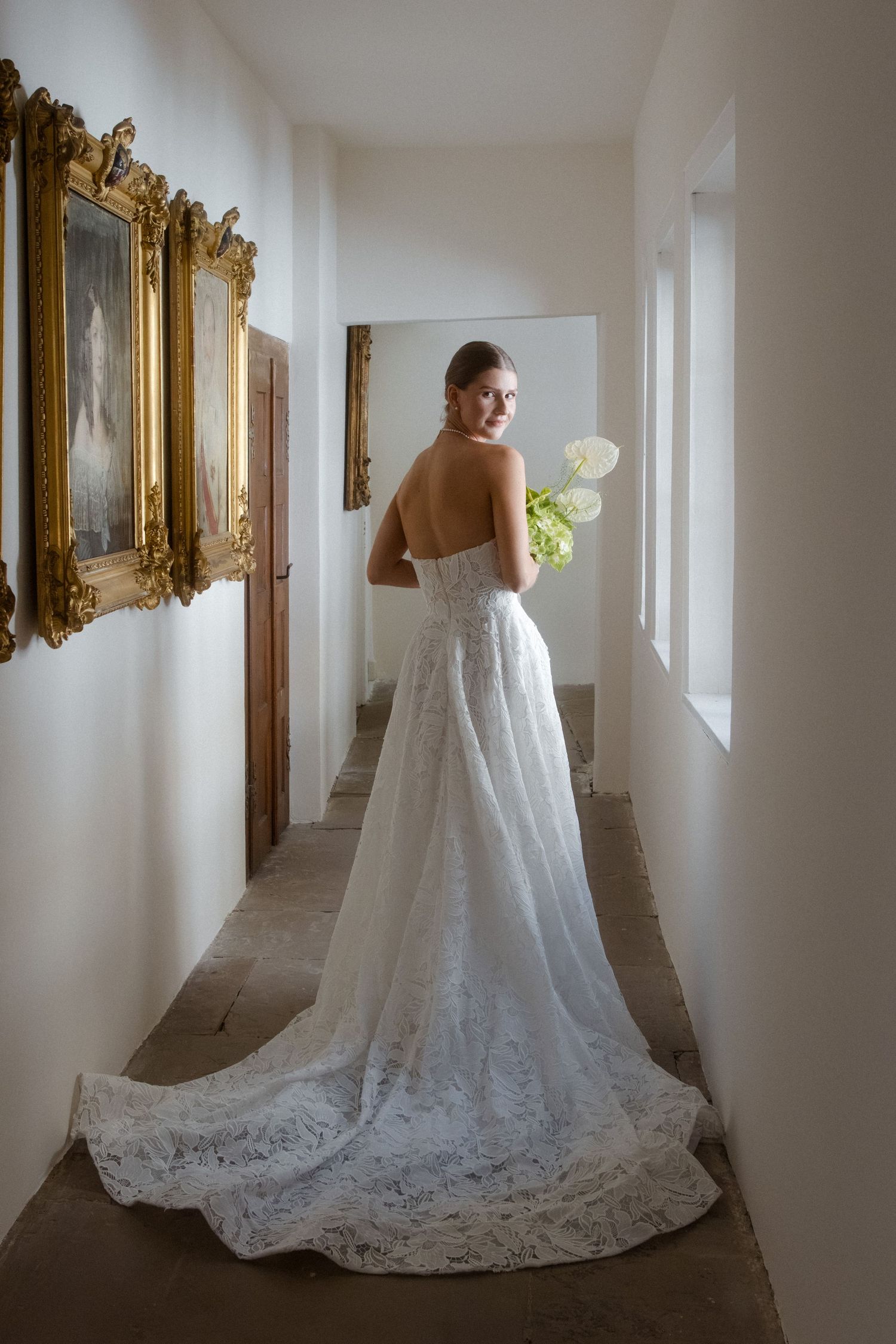 Bride in strapless lace wedding gown with long train holds white bouquet in elegant hallway with gold-framed artwork.