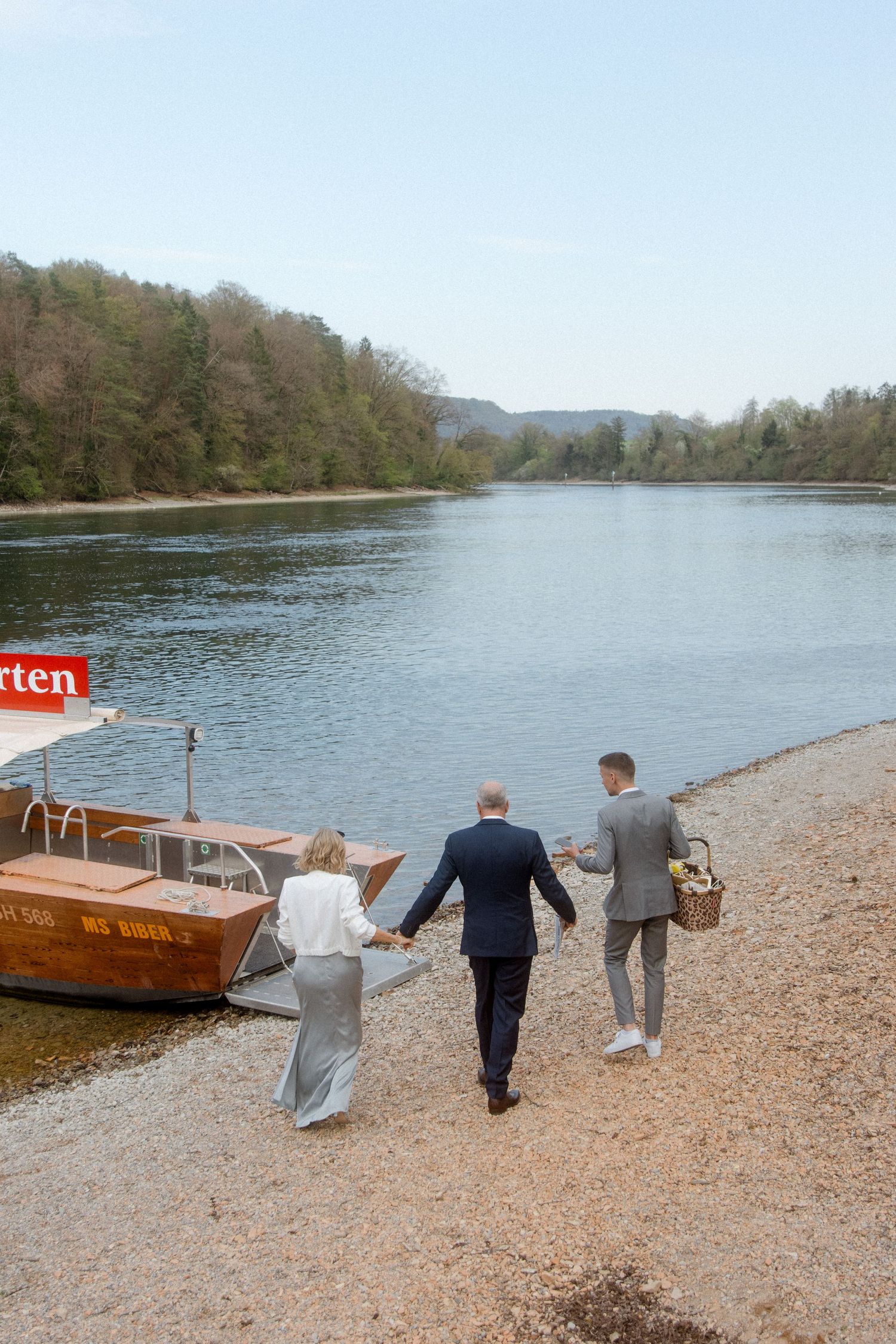 Three elegantly dressed wedding guests walk along a rocky riverbank toward a wooden boat on a calm river.