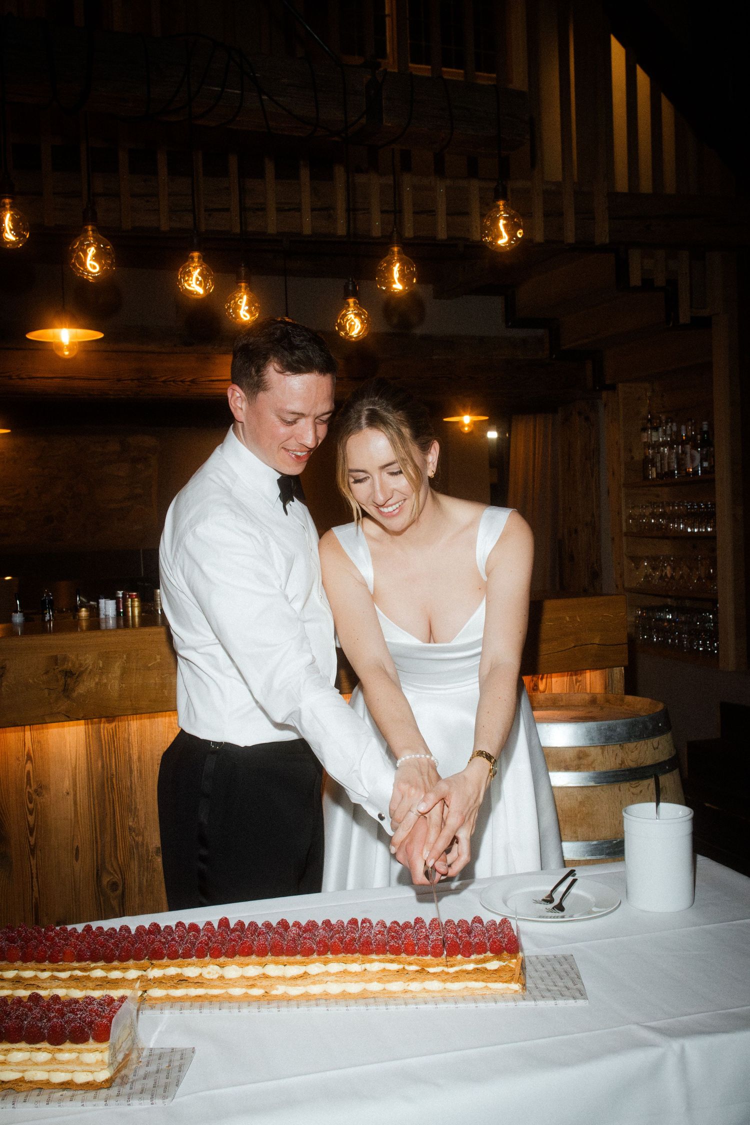 Bride and groom cutting a raspberry mille-feuille wedding cake together in a warmly lit rustic barn venue.