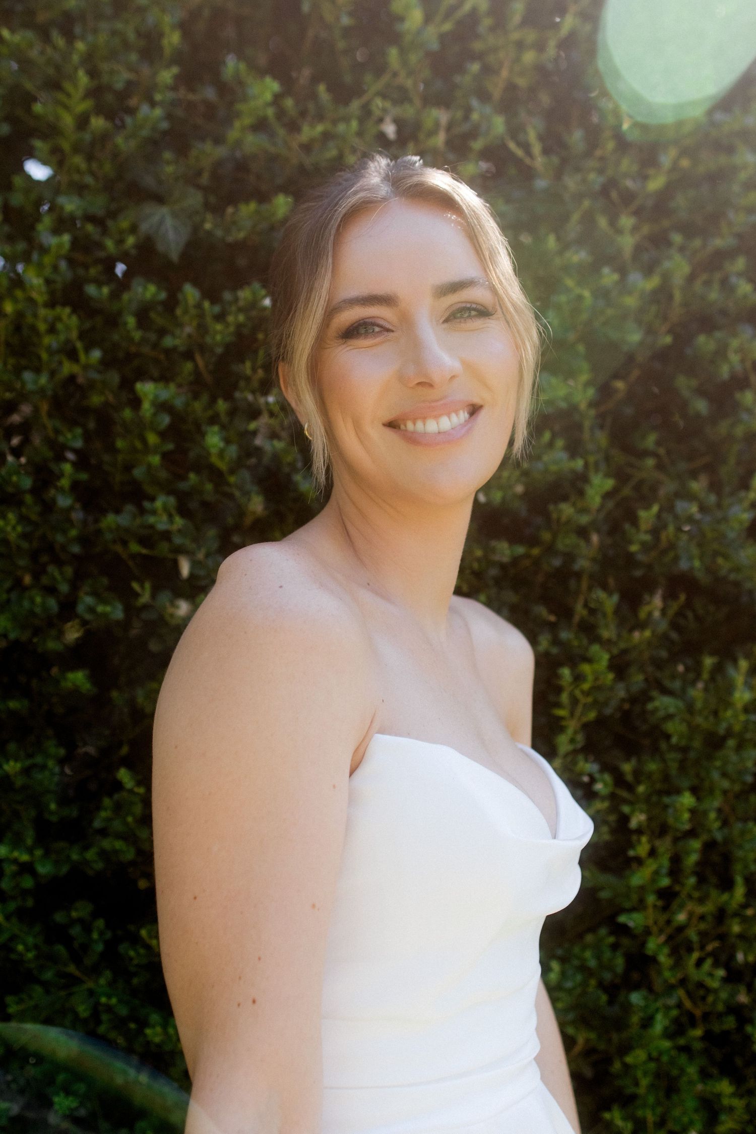 Smiling bride in strapless white gown posing in sunlit garden with green hedges backdrop.
