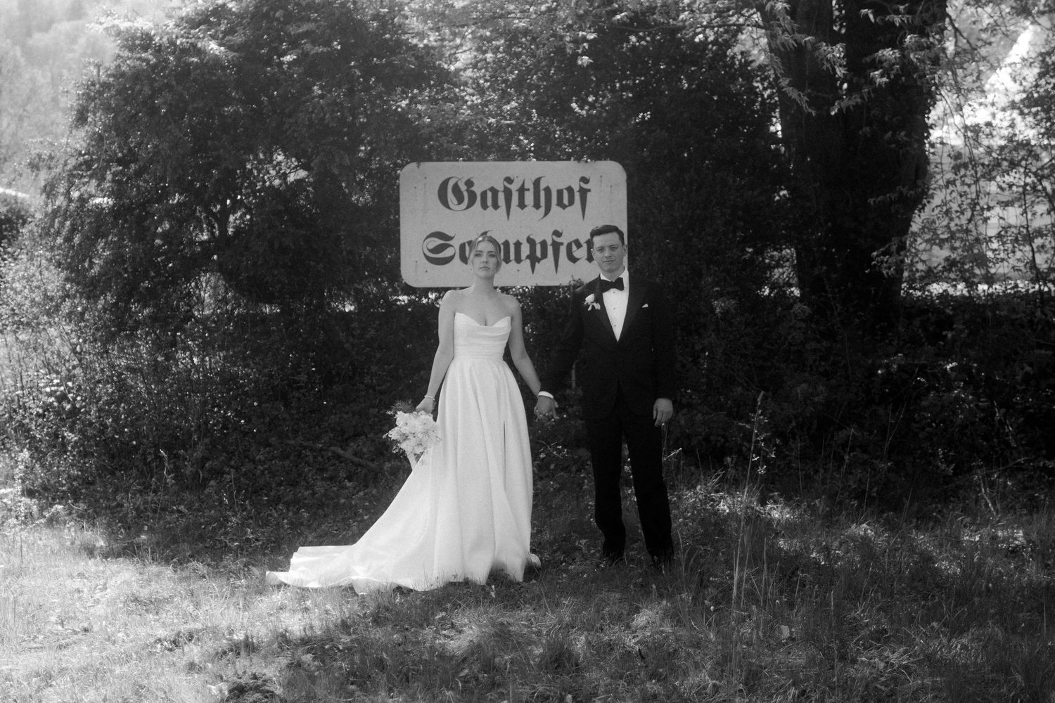 Black and white wedding photo of bride and groom holding hands before a Gasthof Schupfer sign.