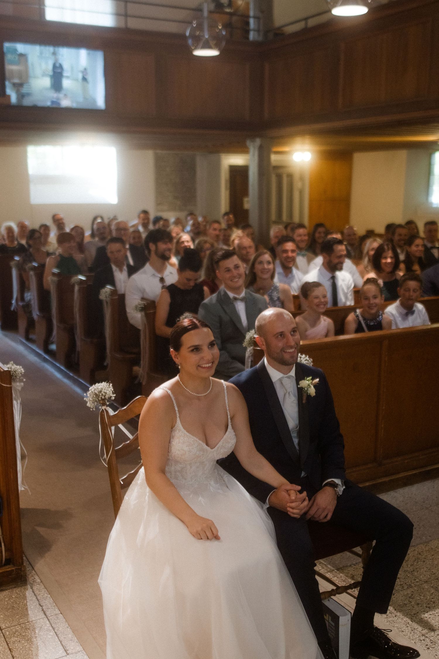 A couple seated in the front of a church during their wedding ceremony with guests visible in background rows.