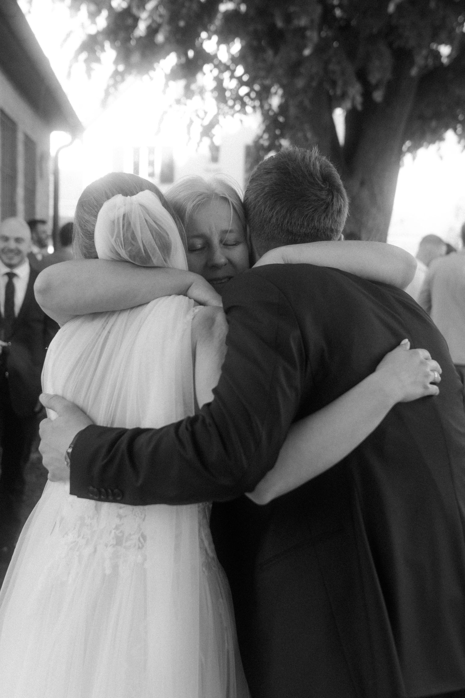 Black and white photo of an emotional group hug between bride, groom and loved one at wedding celebration.