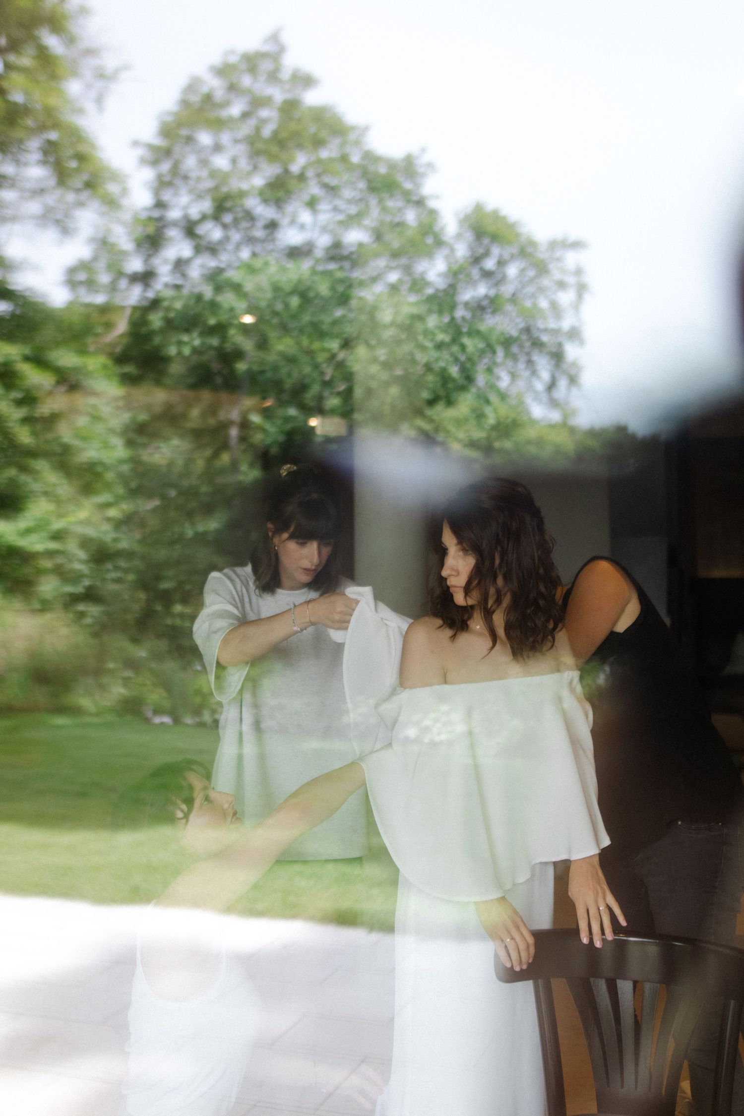 Wedding dress preparation captured through window reflection with green foliage in background.