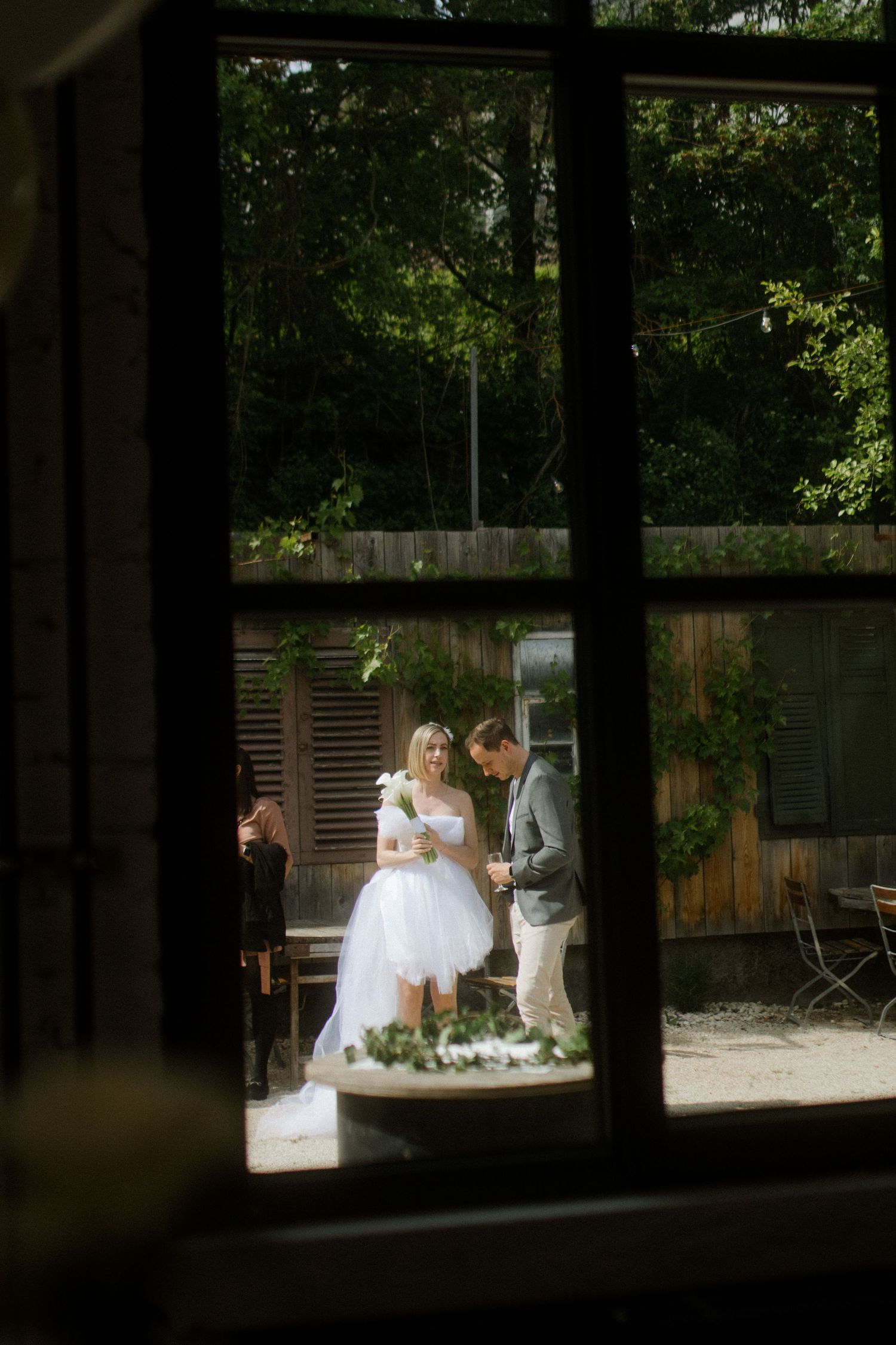 A wedding ceremony viewed through a window frame in a rustic outdoor setting.