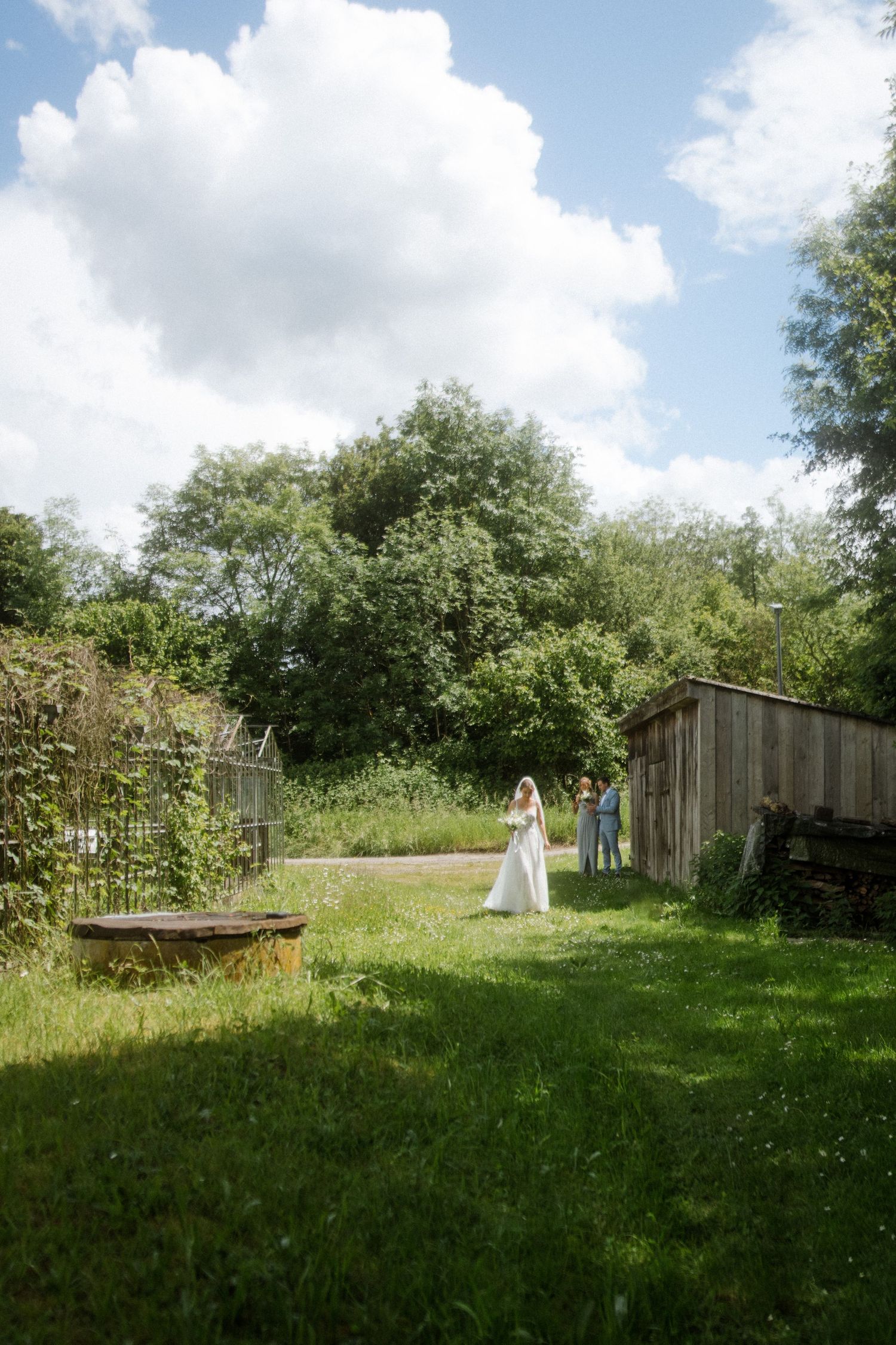 A wedding couple stands in a lush garden setting with wooden fences and trees in the background.