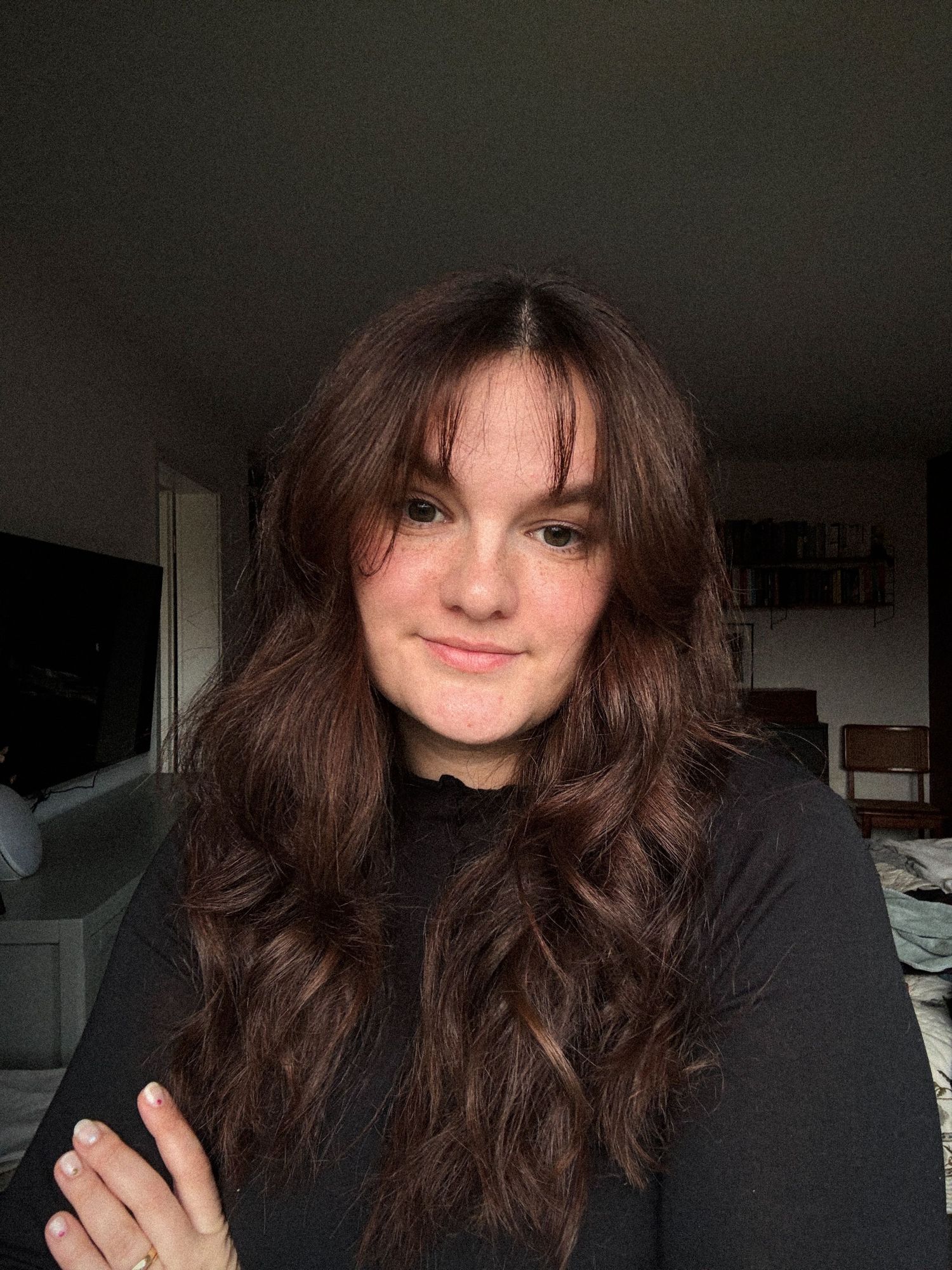 A person with long dark wavy hair wearing a black top poses in a dimly lit room with natural lighting.