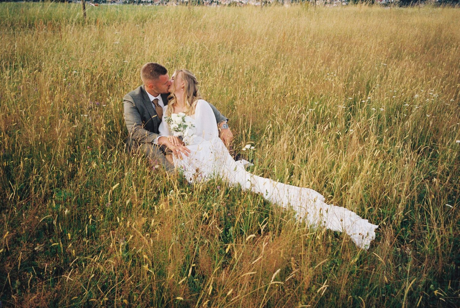 A romantic couple in wedding attire embraces while lying in a golden field of tall grass at sunset.