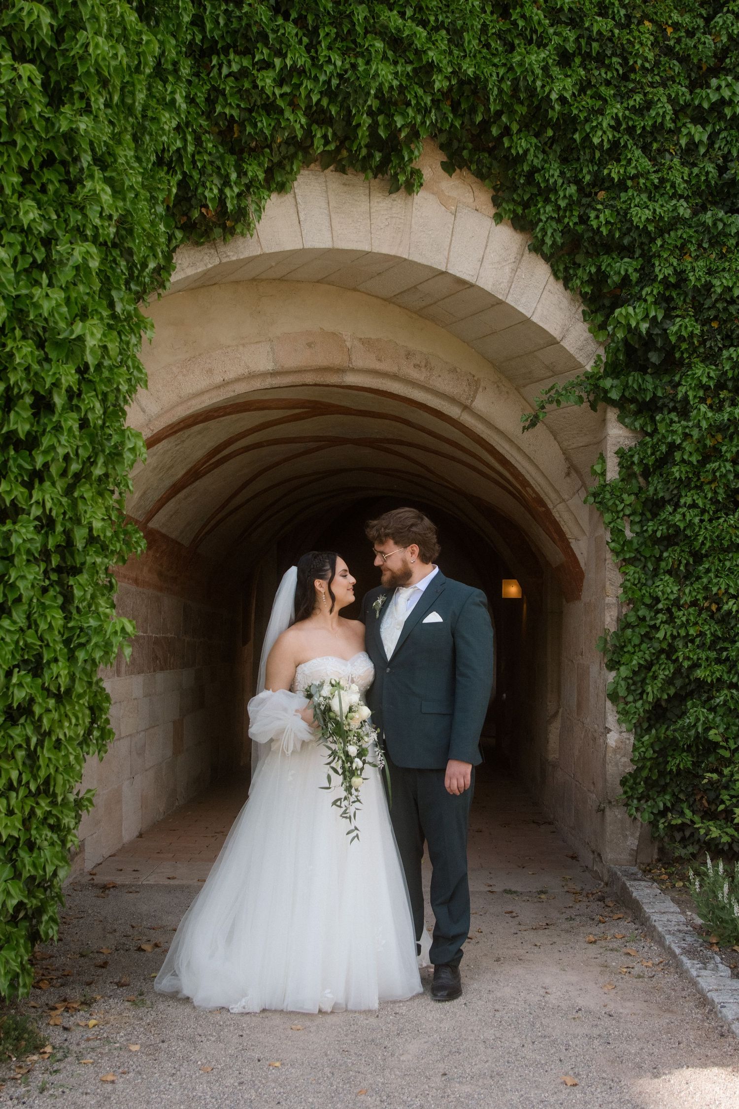 Wedding couple poses in stone archway covered with lush green ivy.