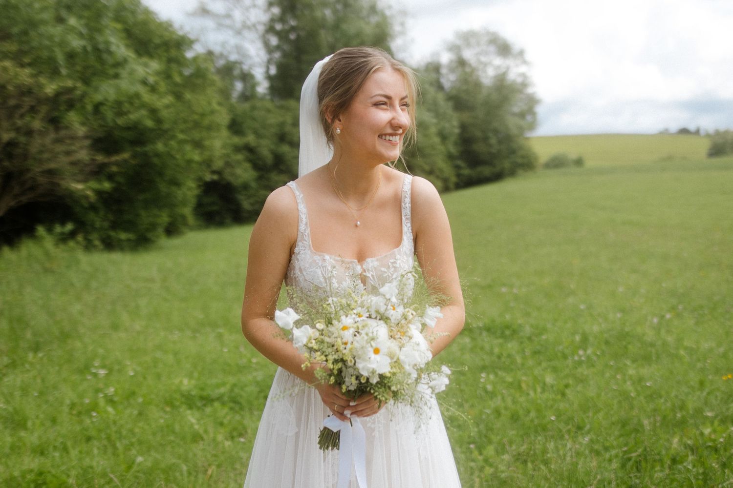 A bride in a white lace dress holds a delicate bouquet of white flowers while standing in a sunny meadow.