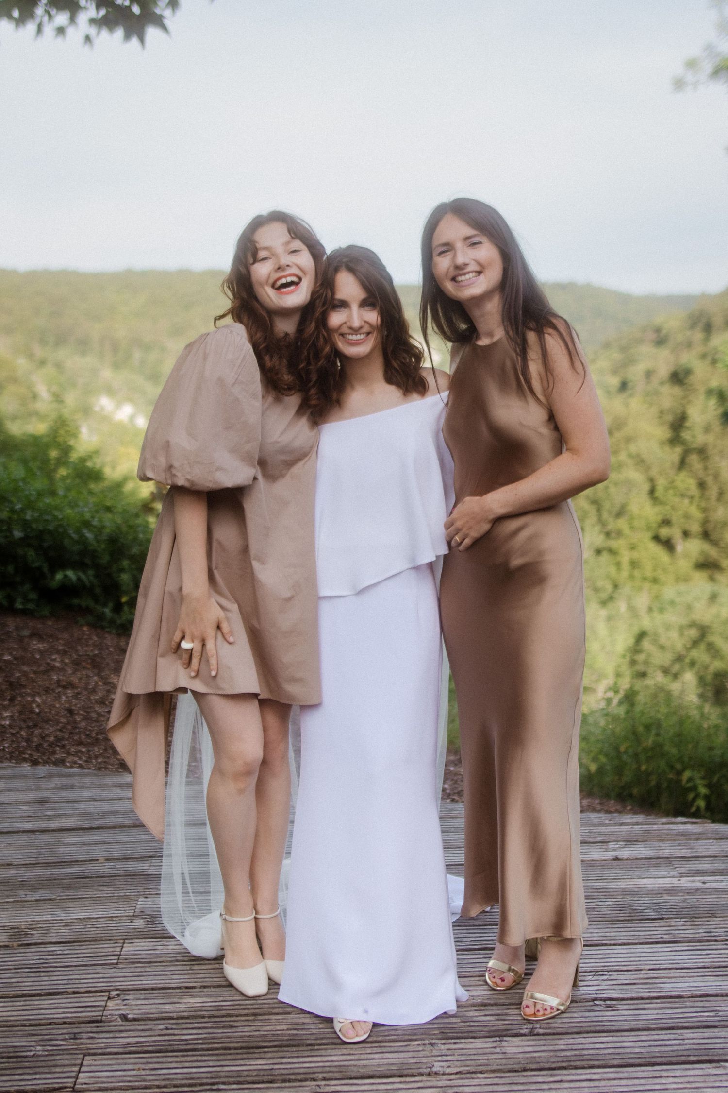 Three women in elegant dresses share a candid moment on a deck with scenic mountain views.