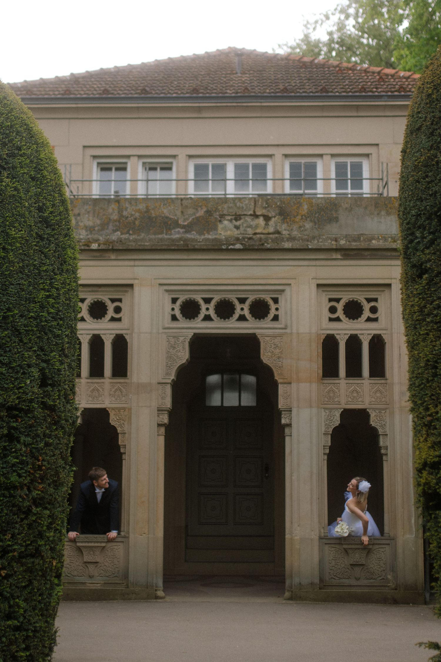 Wedding couple pose in arched doorway of historic building framed by tall hedges.