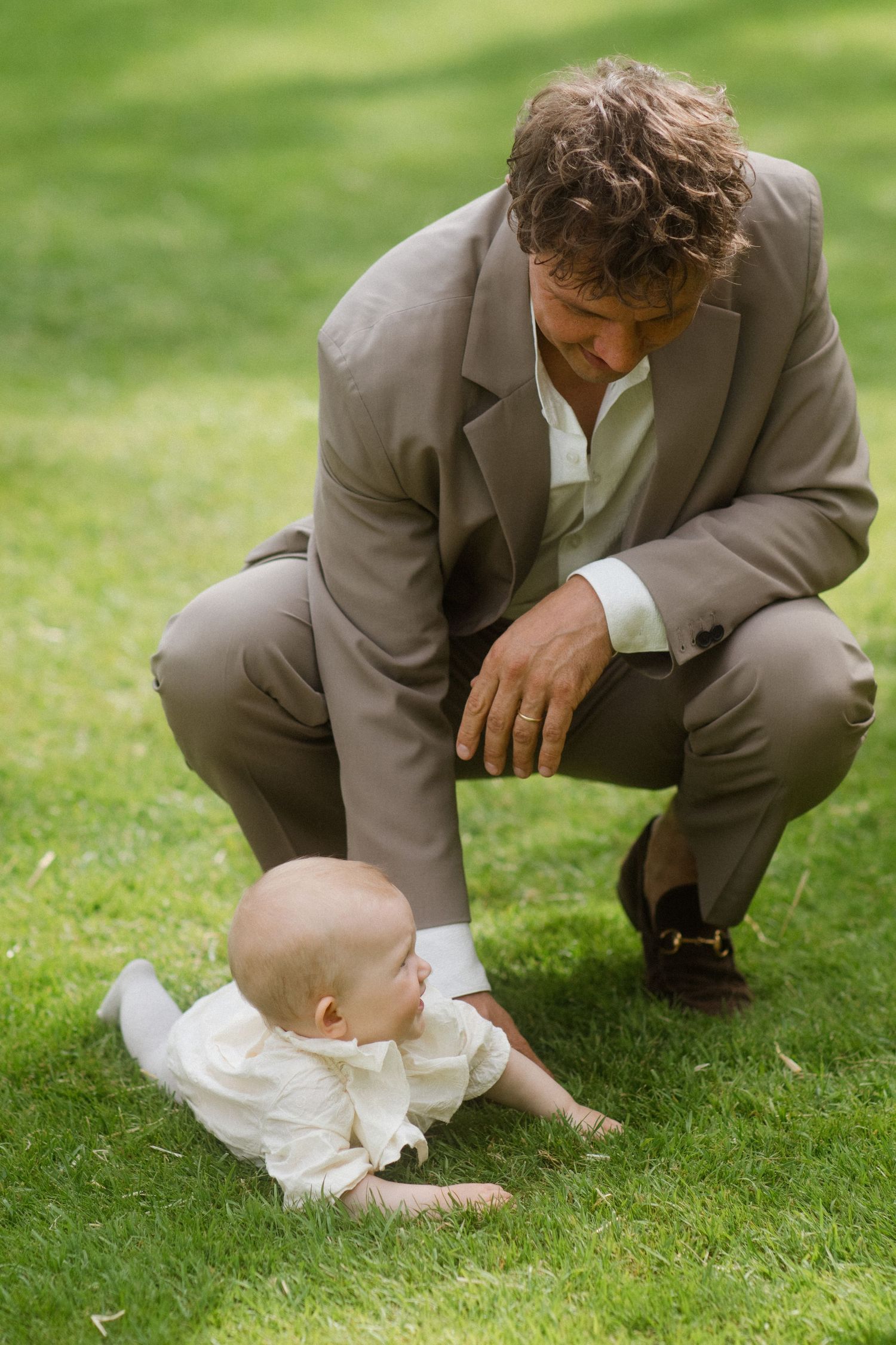 Man in beige suit crouches down to interact with baby on grass.