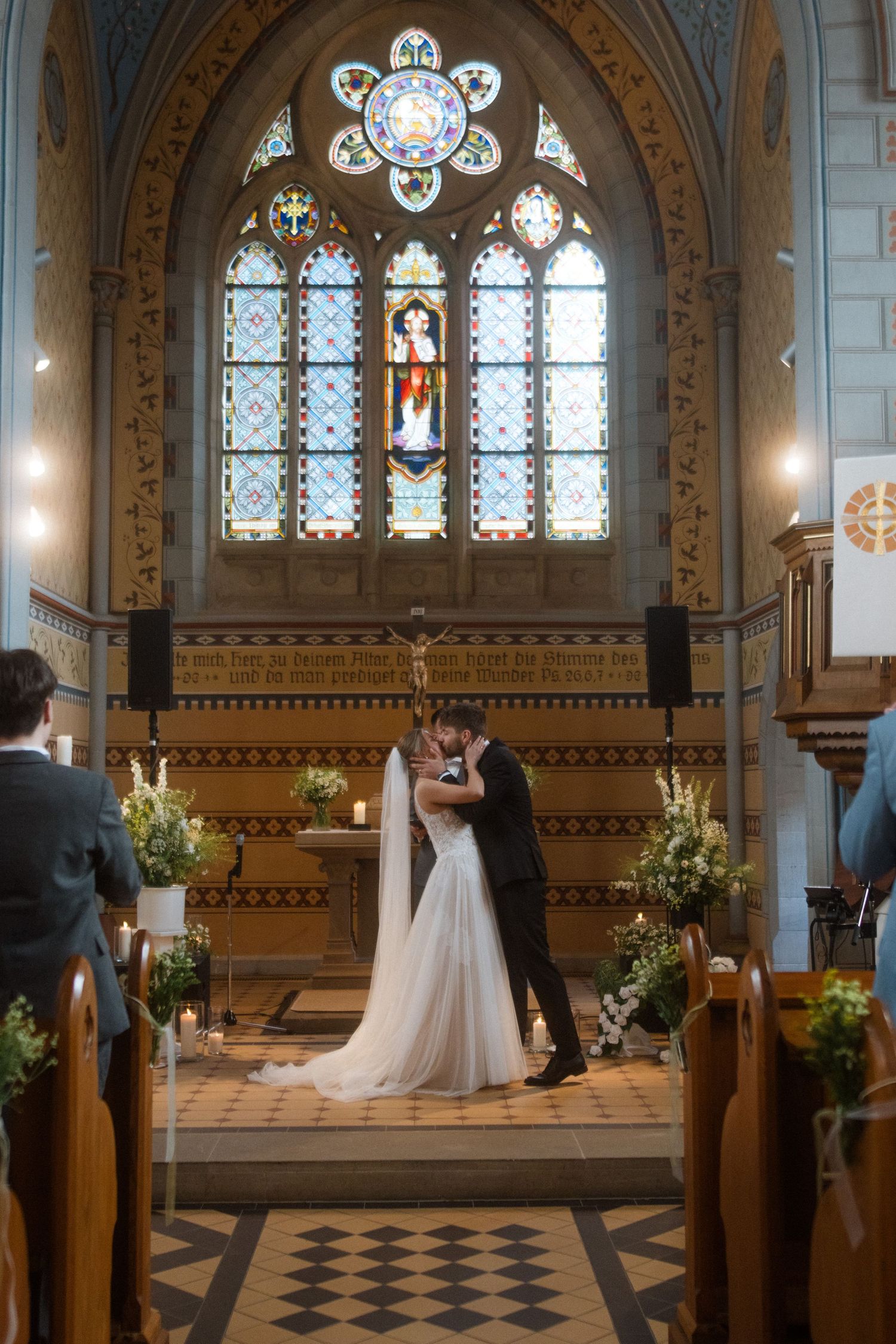 Wedding ceremony in church with ornate stained glass windows and floral decorations.