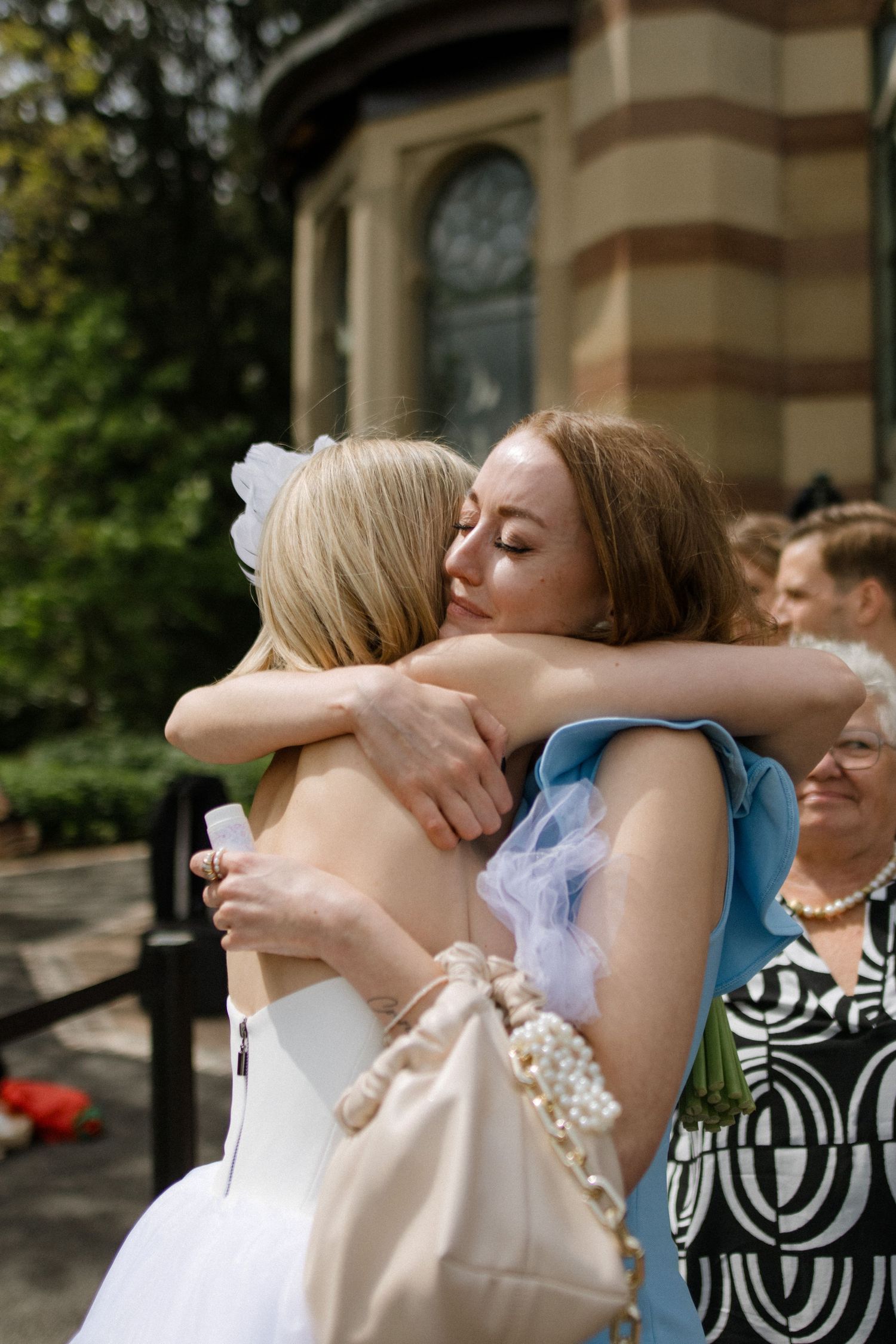 Two wedding guests share a heartfelt embrace outside a historic brick building.