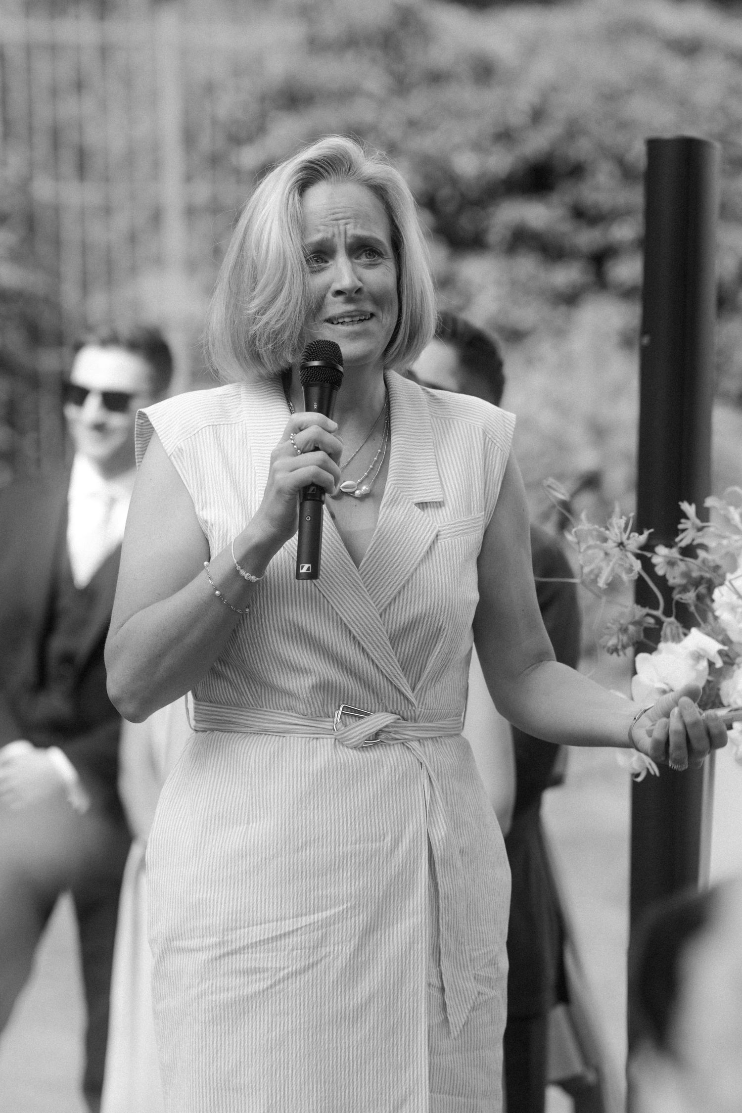 Speaker in white dress addresses audience at outdoor gathering.