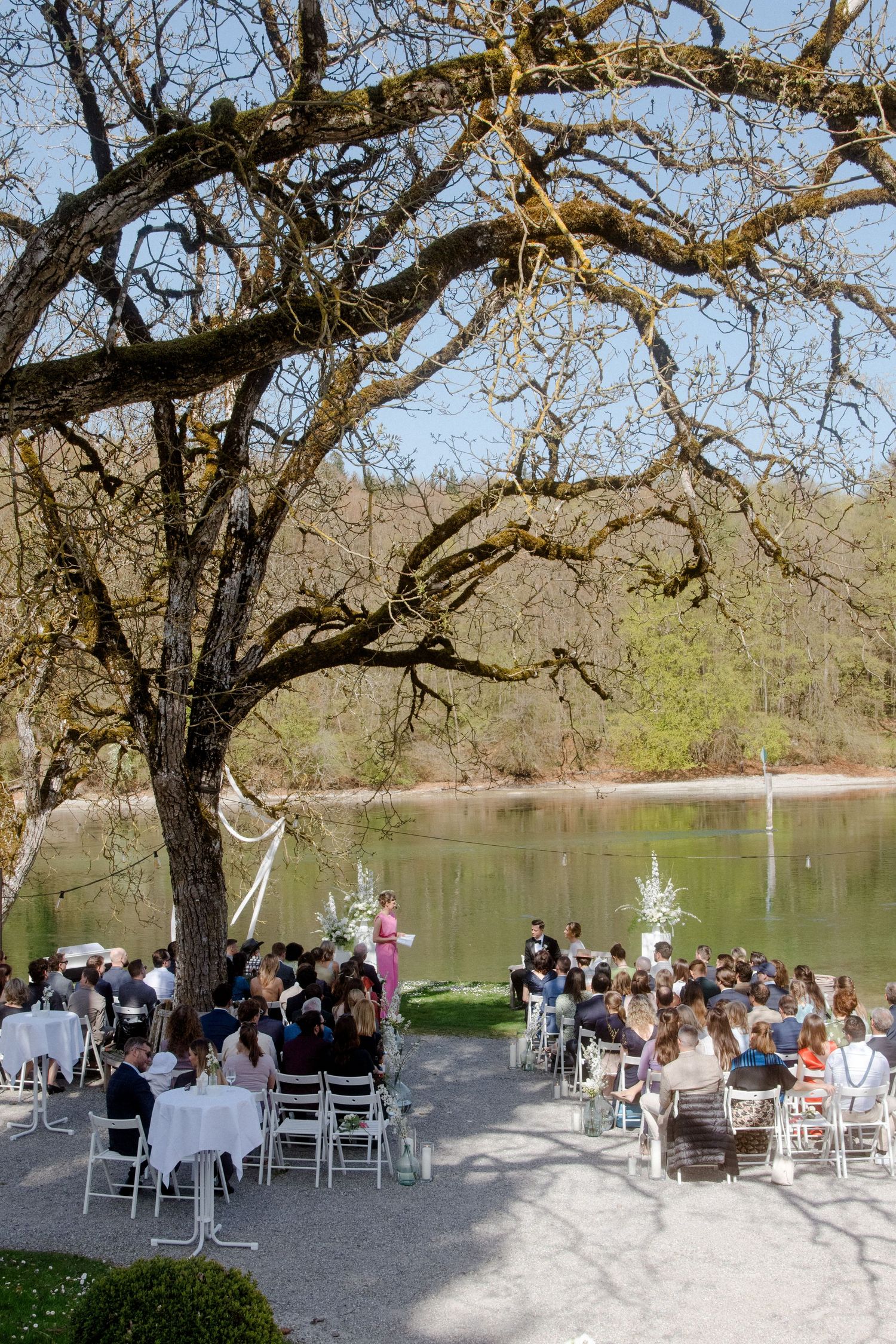 Outdoor wedding ceremony beside a serene lake, guests seated on white chairs beneath a large bare tree.