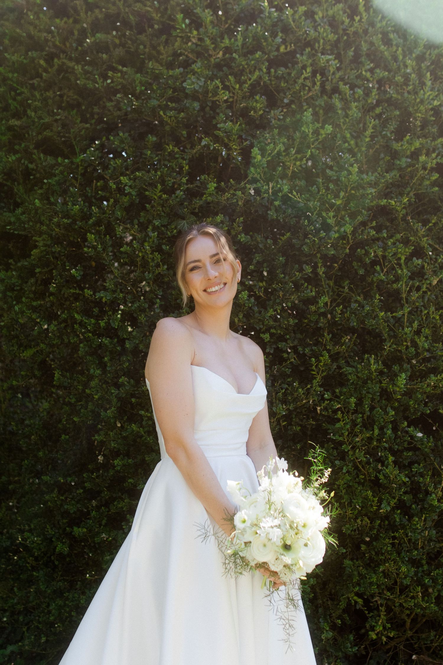 Smiling bride in white strapless gown holding white bouquet, posing in front of lush green hedge in sunlight.