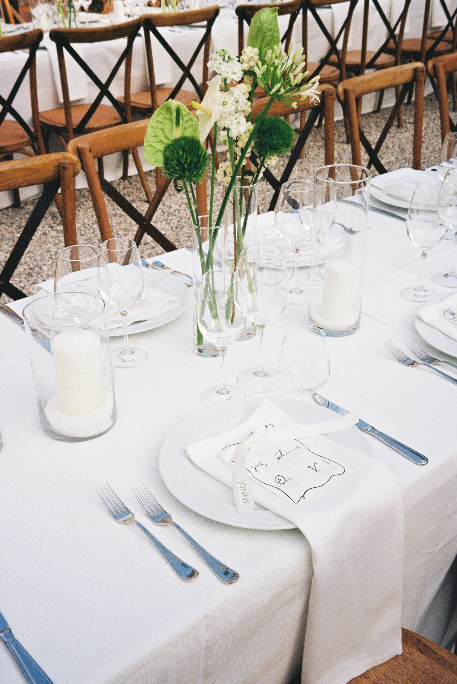 Elegant white table settings with wooden chairs and simple floral centerpieces at an outdoor reception.