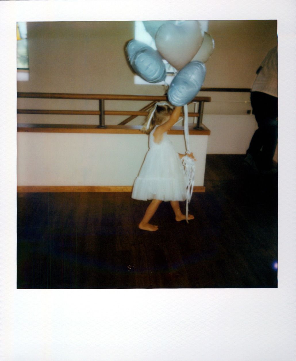 A dancer in a white dress holds balloons while practicing in a dance studio with wooden floors and ballet barre.