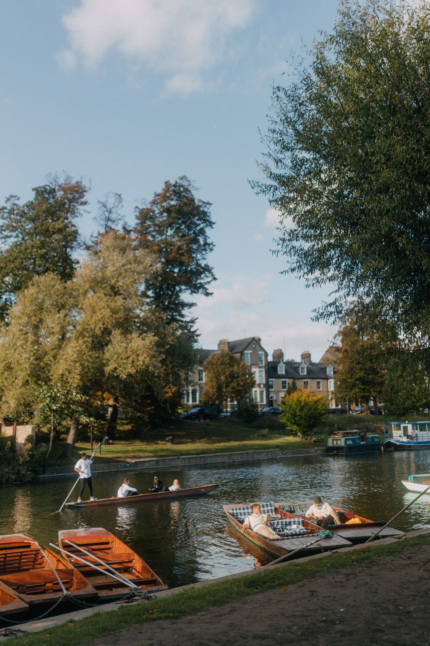 Wooden boats line the shore of a tranquil lake with houses visible in the background on a sunny autumn day.