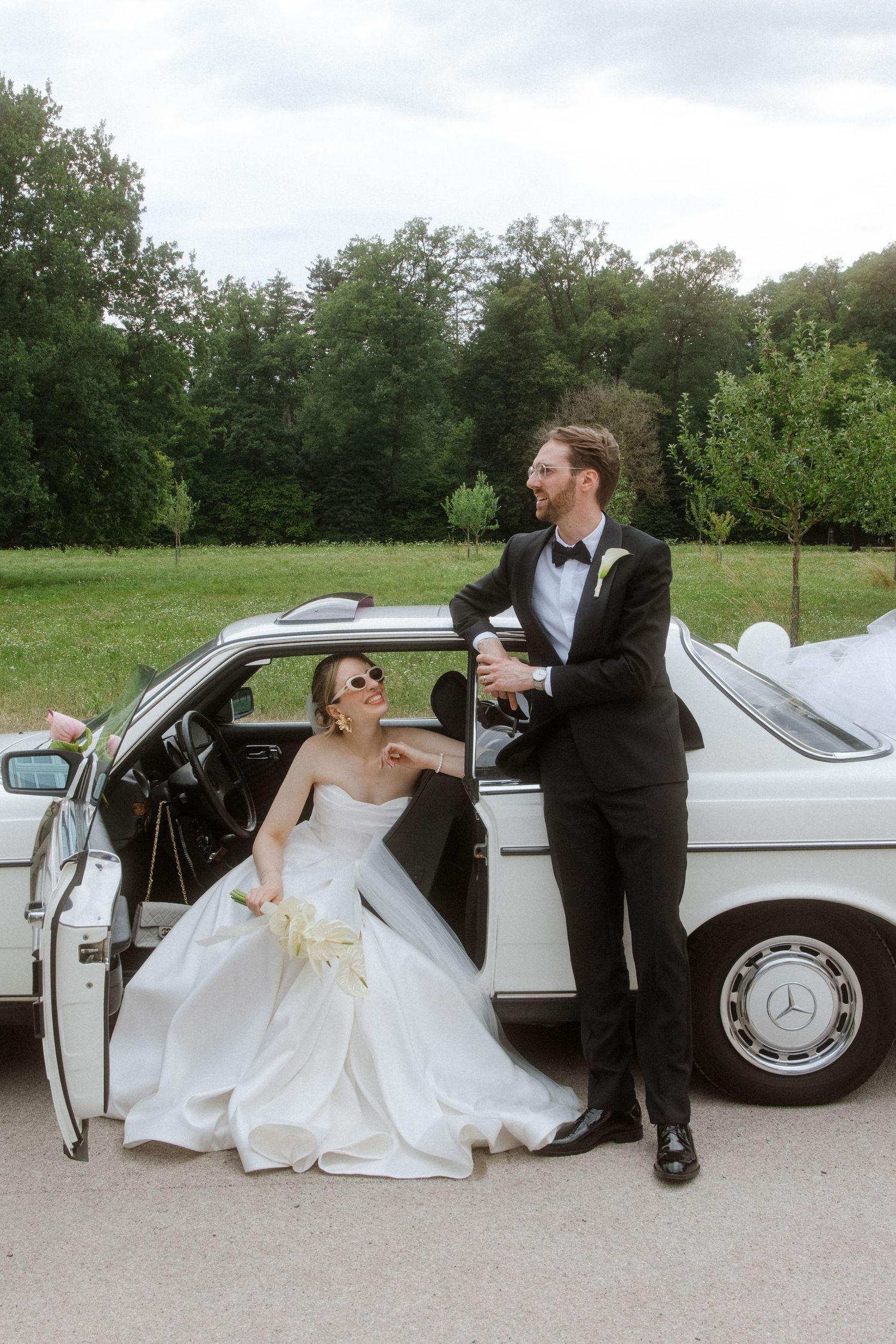 Wedding couple poses playfully by classic white car in outdoor setting.
