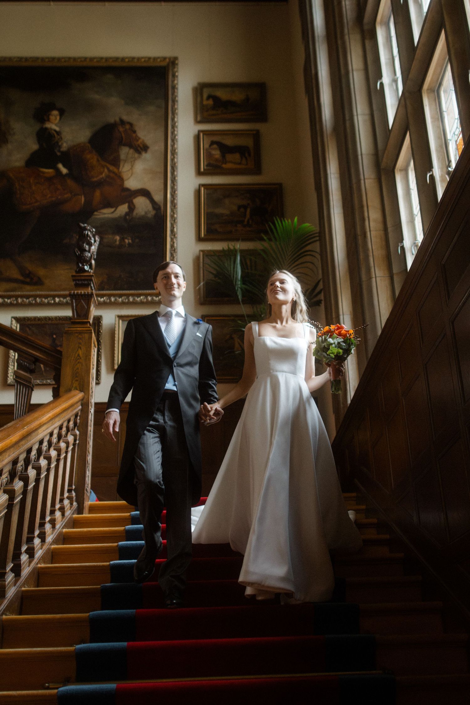 A couple descends a grand staircase decorated with equestrian paintings and wooden paneling.