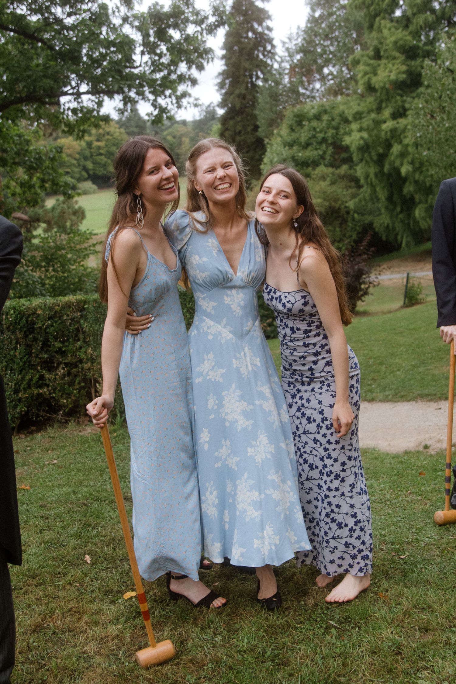 Three friends in summer dresses laughing together on a grassy lawn during a sunny day.