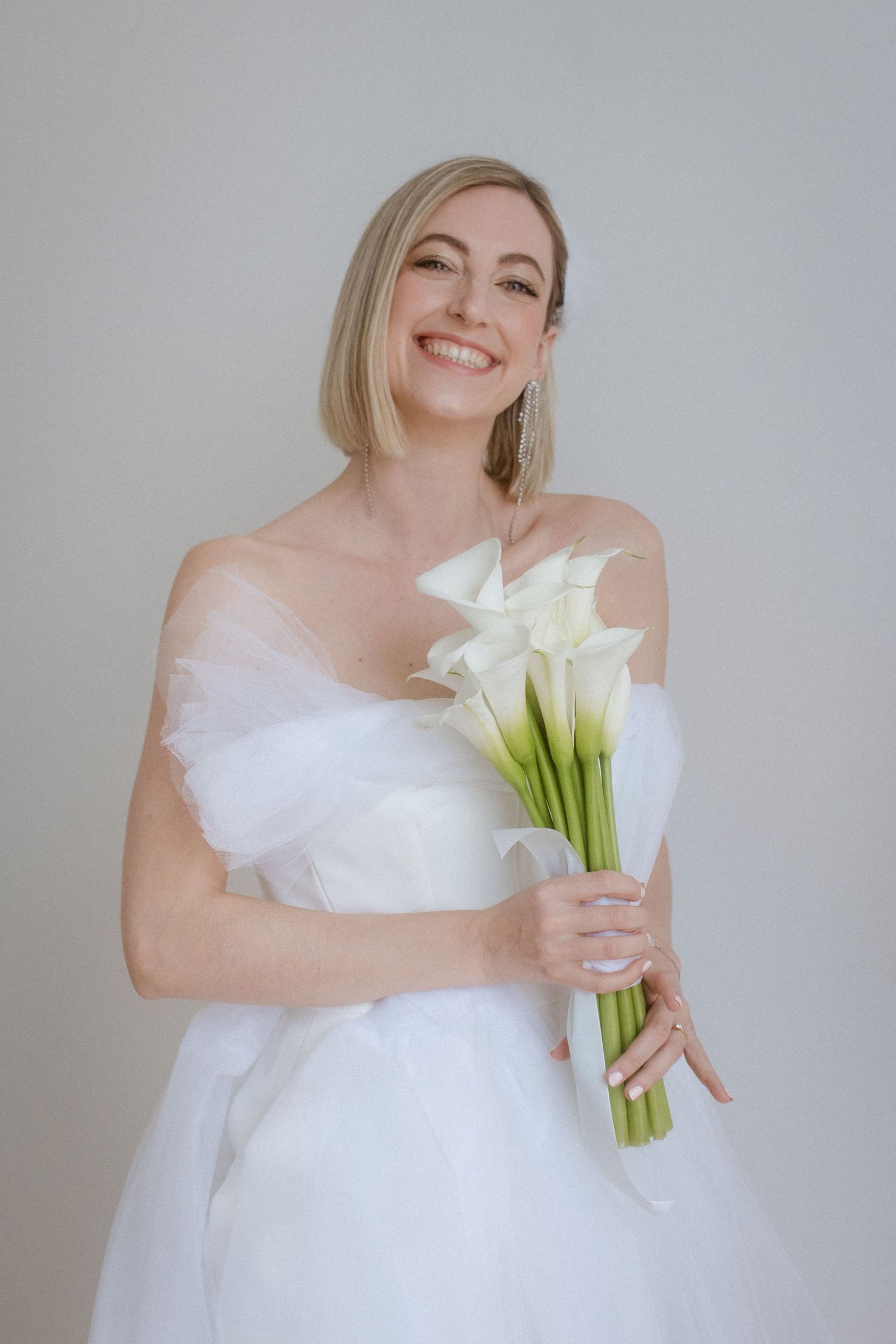 Joyful bridal portrait with white calla lily bouquet against neutral background.