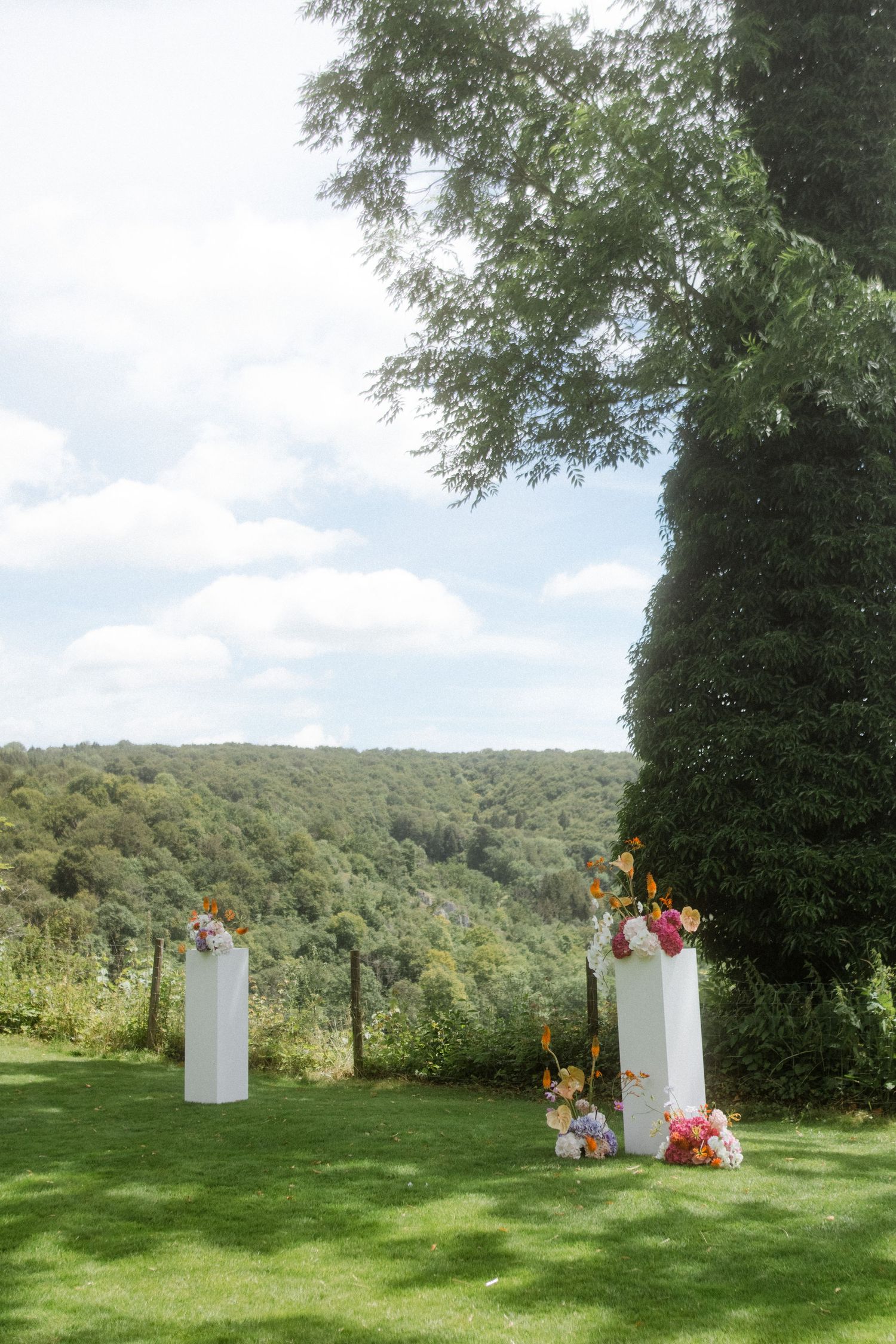 Two white ceremony pillars with floral arrangements set against scenic hillside landscape.