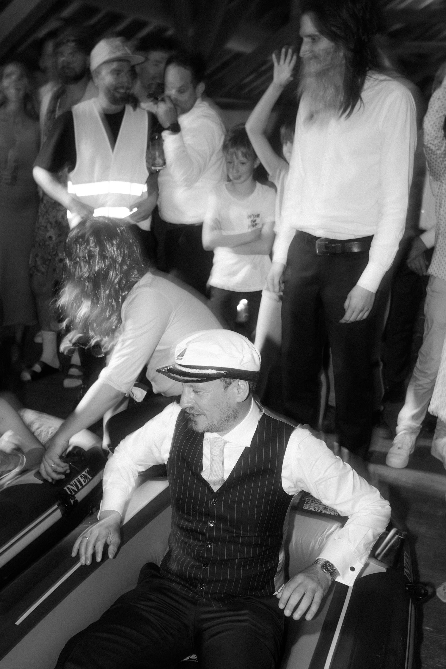 Man in vintage attire and flat cap sits at party while others gather around in black and white photo.