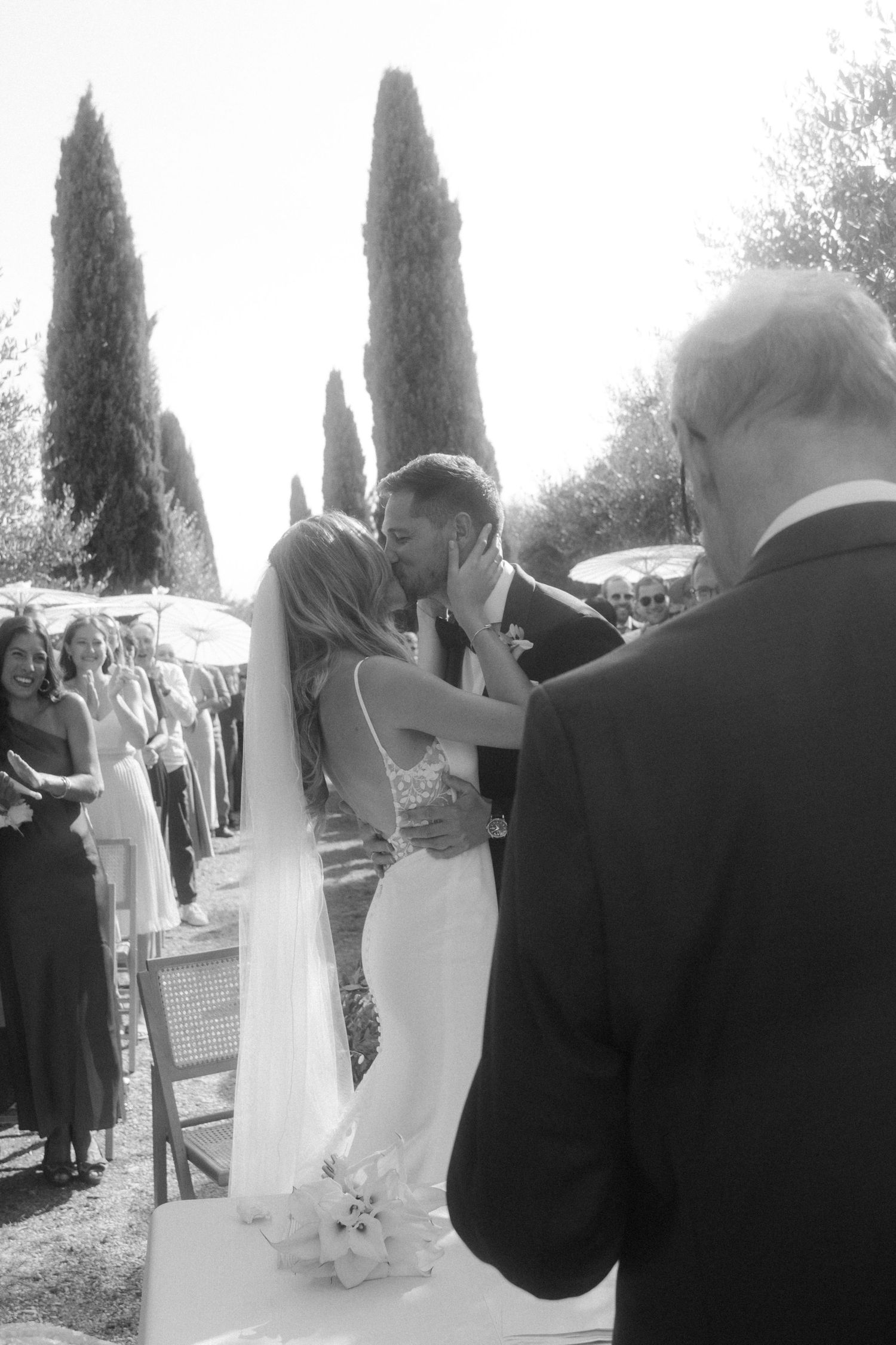 Romantic wedding kiss moment framed by tall cypress trees in black and white.