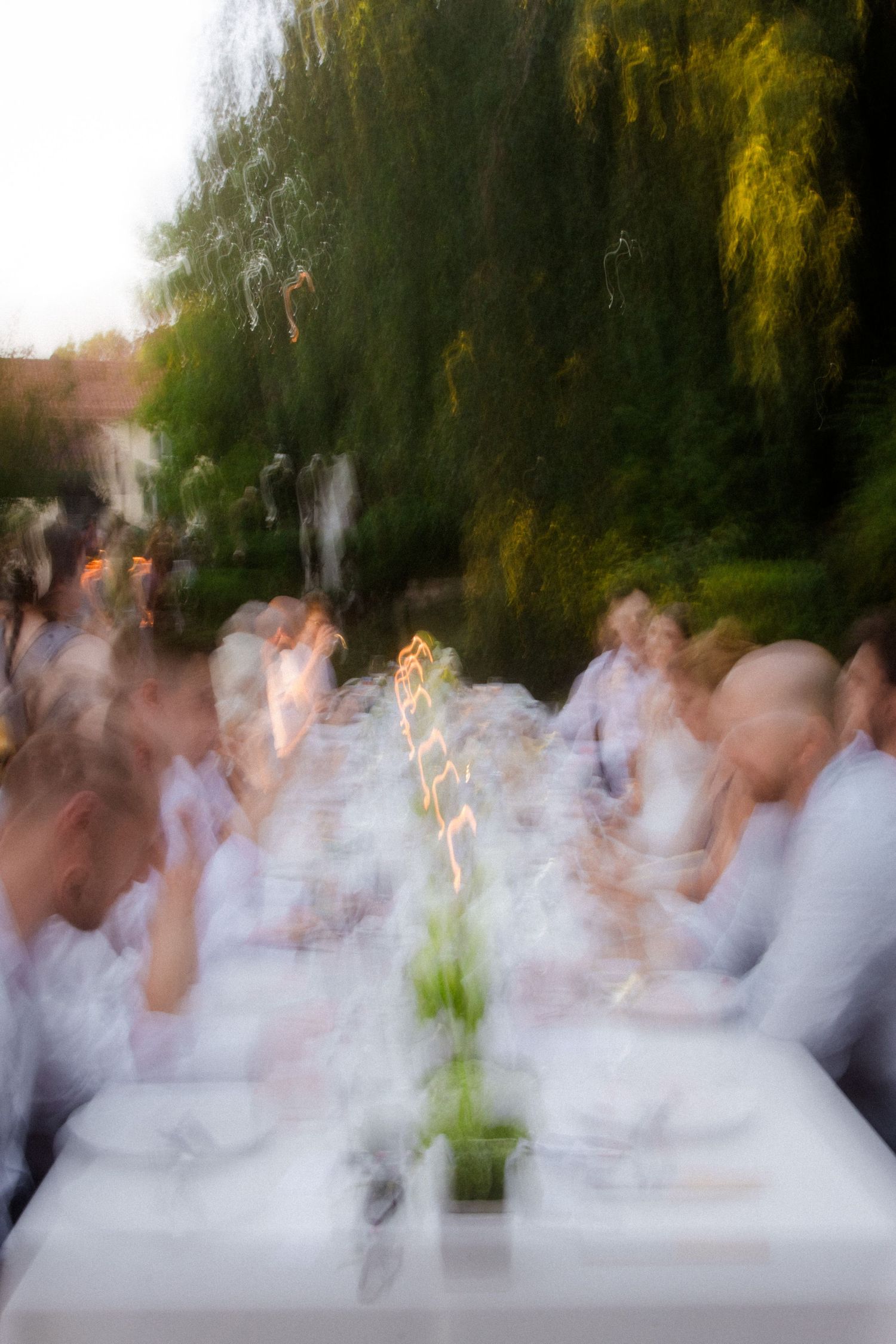 Blurred motion photo of guests in white at an elegant outdoor candlelit dinner party surrounded by lush green trees.