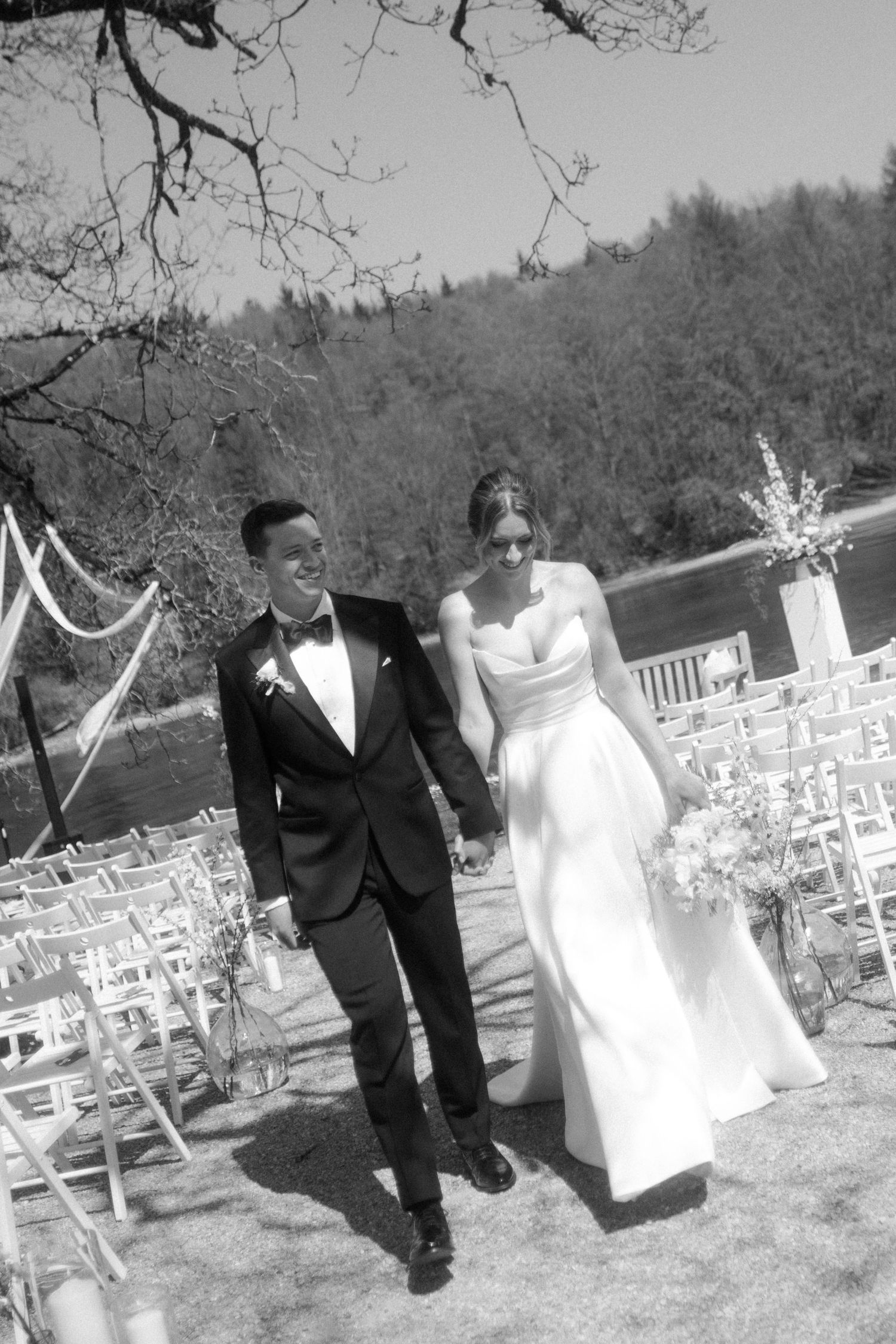 Black and white photo of a bride and groom walking hand in hand down the aisle at an outdoor wedding ceremony.