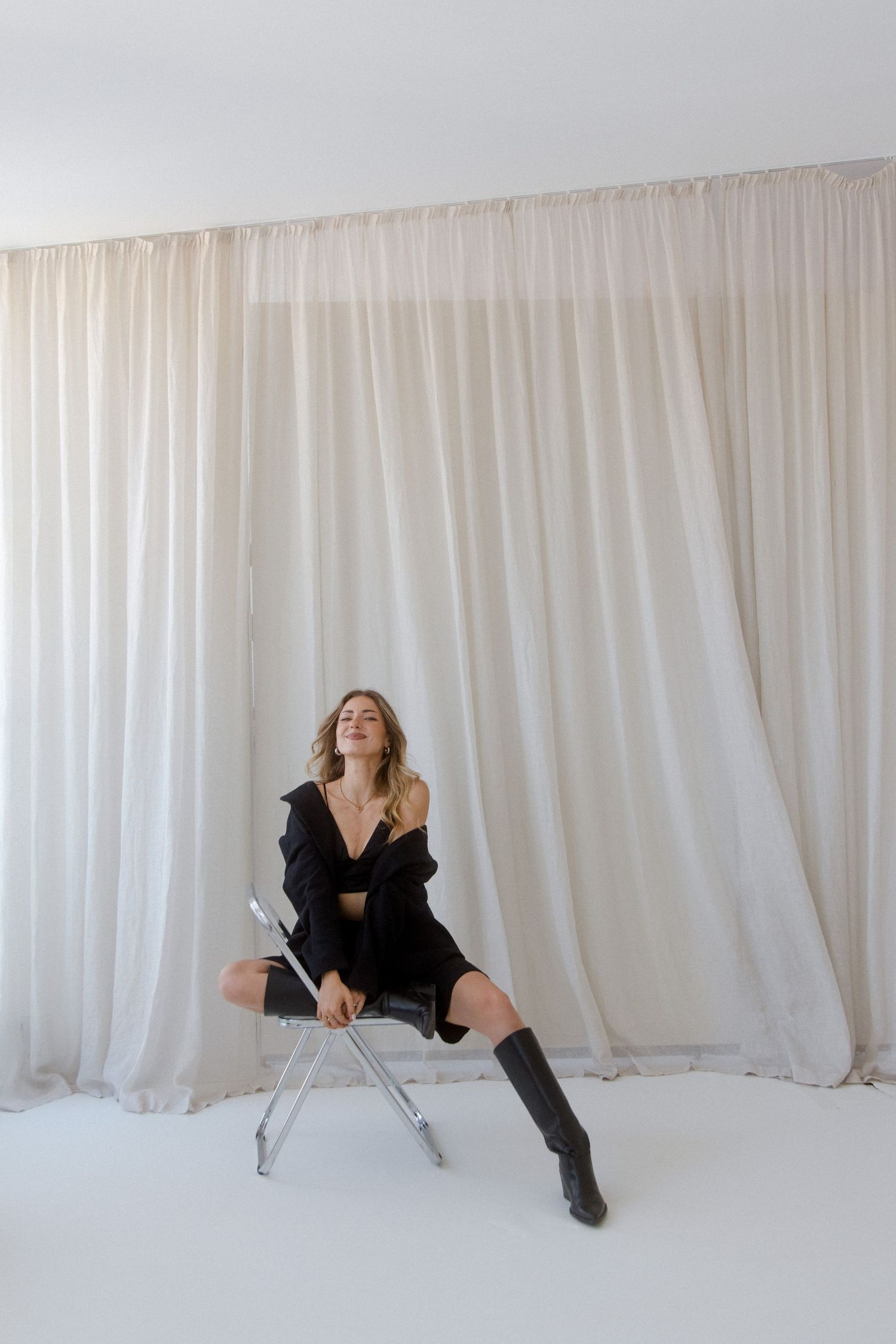 Woman in black outfit and knee-high boots seated on a clear chair against white curtain backdrop.