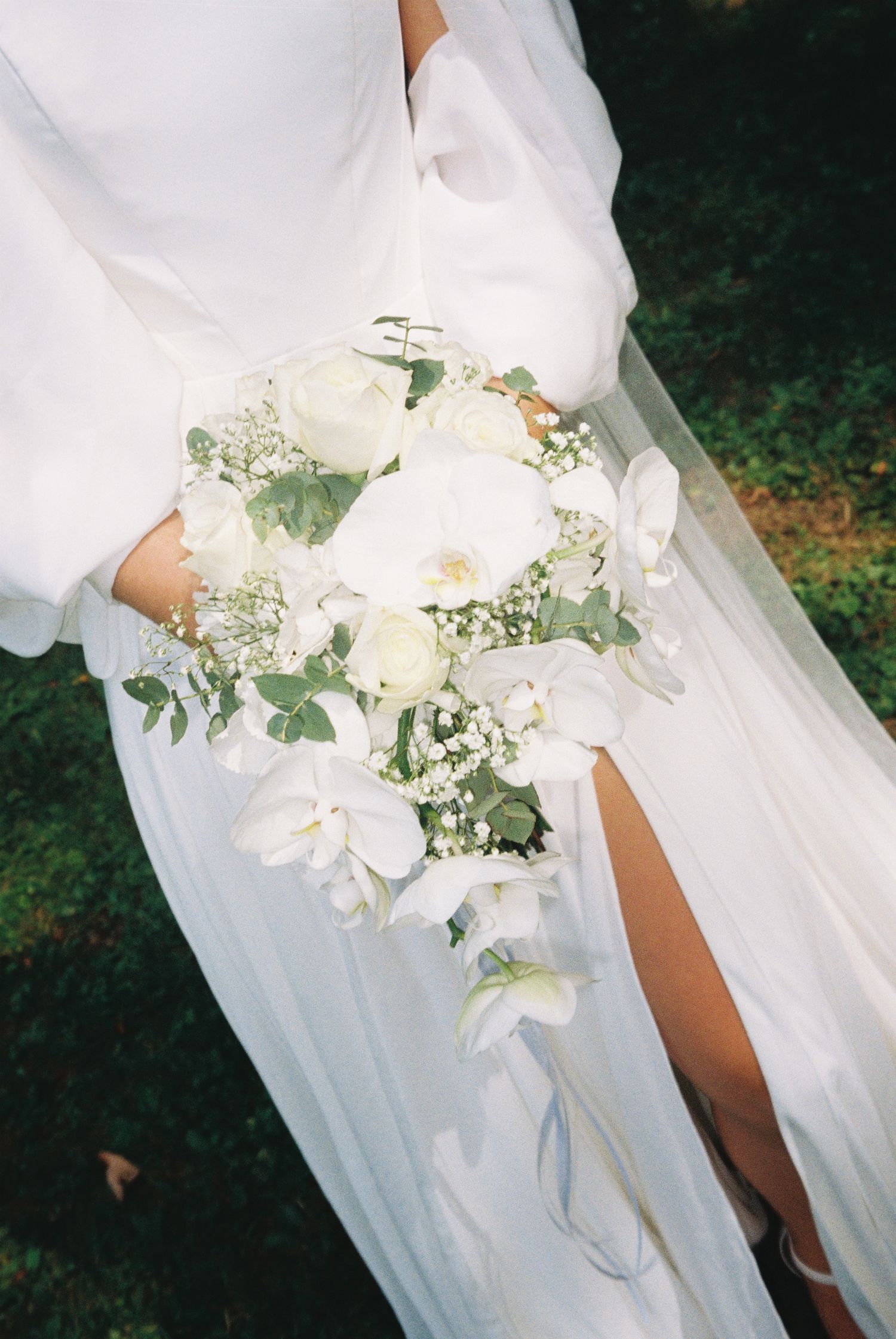 Bride in white gown holding cascading bouquet of white orchids, roses, and baby's breath on wedding day.