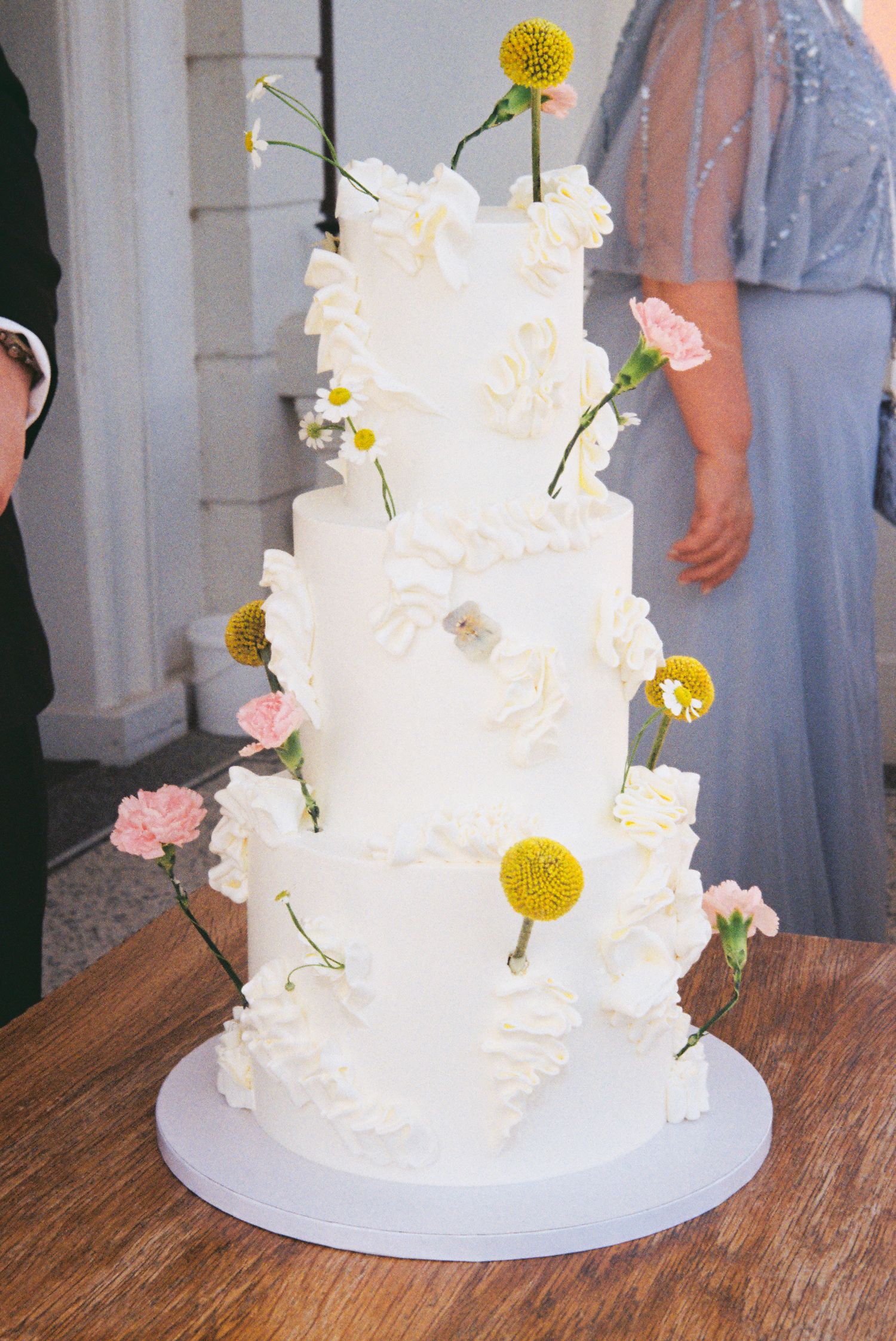 Three-tier white wedding cake with textured frosting, pink carnations, yellow billy balls, and daisy flowers.