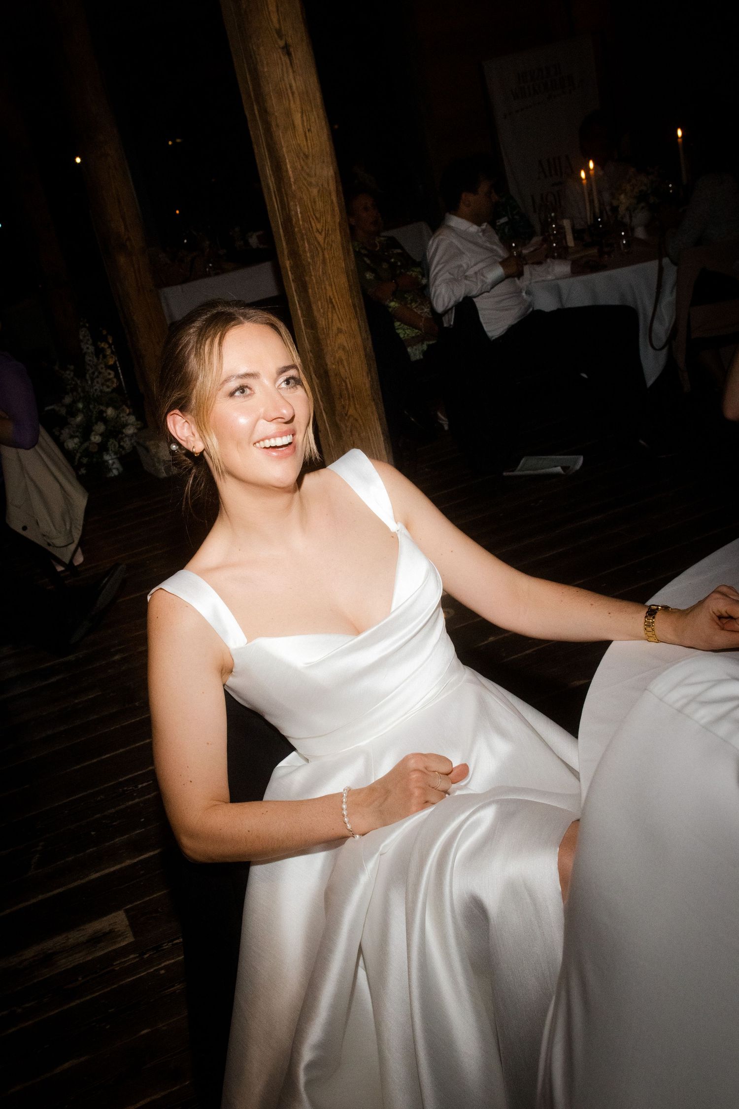 Smiling bride in a white satin gown seated at a candlelit wedding reception in a rustic barn venue.
