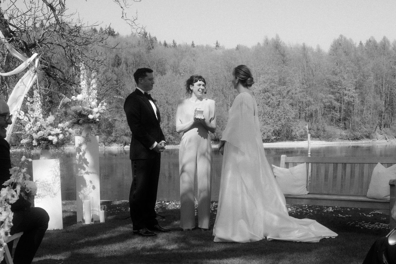 Black and white outdoor wedding ceremony with couple exchanging vows before officiant, lakeside forest backdrop.